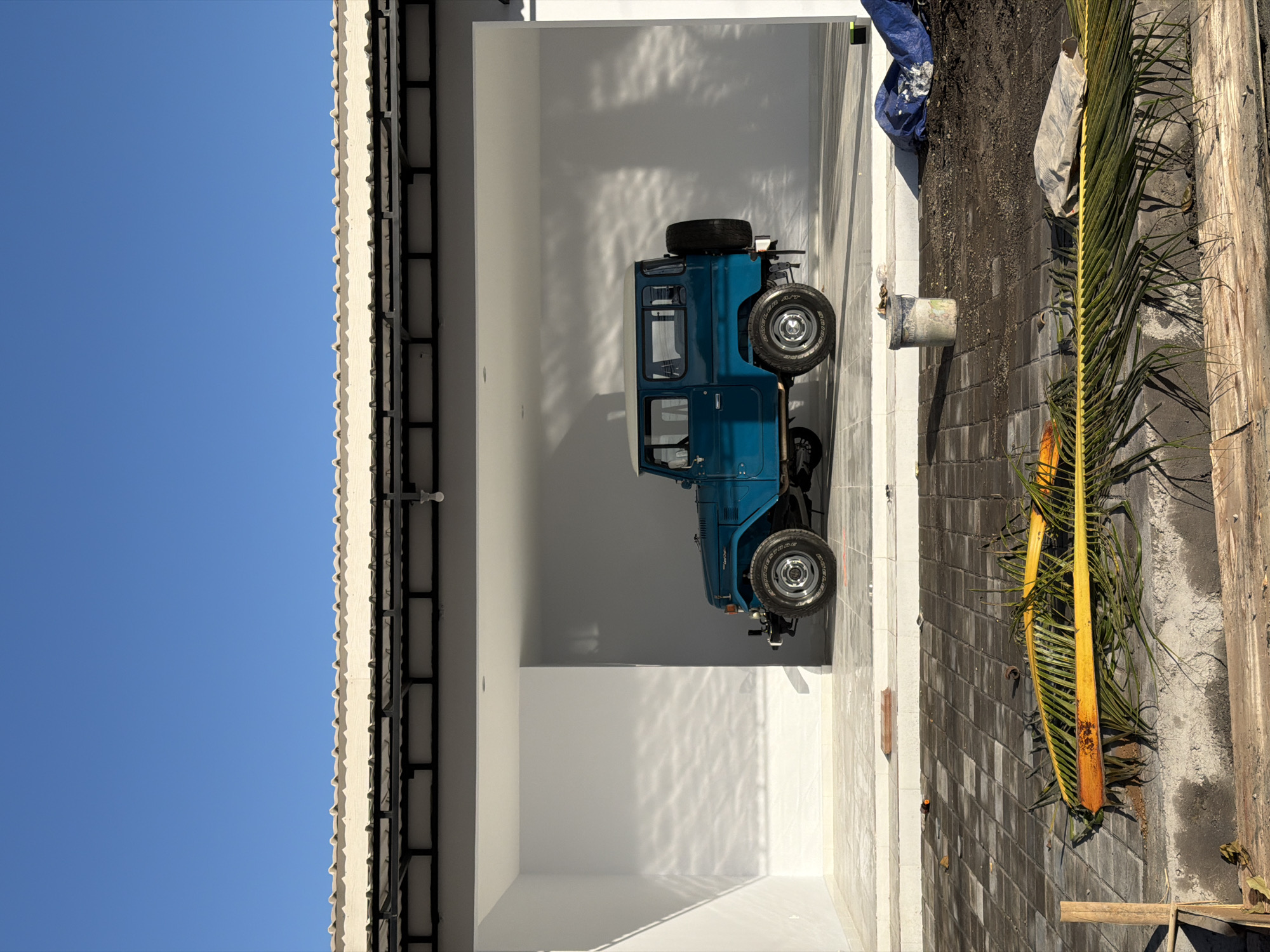 The image, taken in what appears to be North Kuta, Indonesia, shows a teal-colored vintage Toyota Land Cruiser parked inside a freshly painted white garage. The garage is a simple, enclosed space with no visible clutter beyond the vehicle. Outside the garage, there is a brick-paved walkway with some debris and palm fronds scattered around. A partially visible blue tarp is also present near the garage entrance. The sky is clear and blue, suggesting a sunny day. The image suggests an ongoing construction or renovation project.