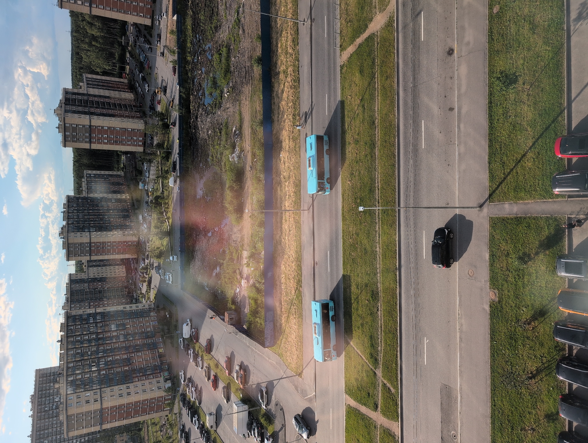The image shows a high-rise residential area in Saint Petersburg, Russia, with several tall apartment buildings visible. The foreground features a road with two light blue buses driving on it. A black car is parked on the side of the road. A patch of grass is on the left side of the road. In the background, more apartment buildings and a forest area are visible. The sky is partly cloudy.