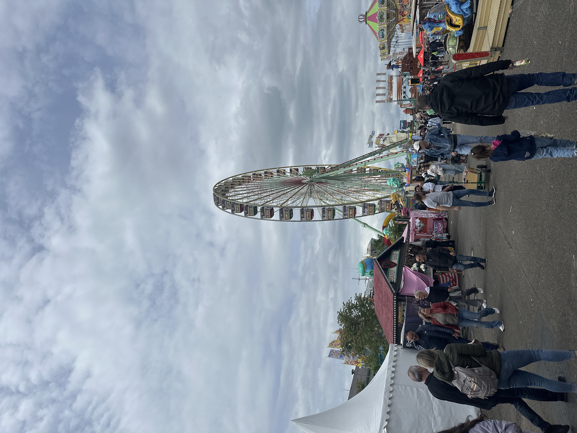 The image depicts a scene at a fair or carnival in Heidelberg, Germany. The sky is overcast with varying cloud formations. A large Ferris wheel dominates the view, with its frame and carriages visible. There are numerous people walking through the fair, suggesting a lively atmosphere. Food stalls and carnival rides are set up along the sides of the path. A large white tent is partially visible in the foreground.