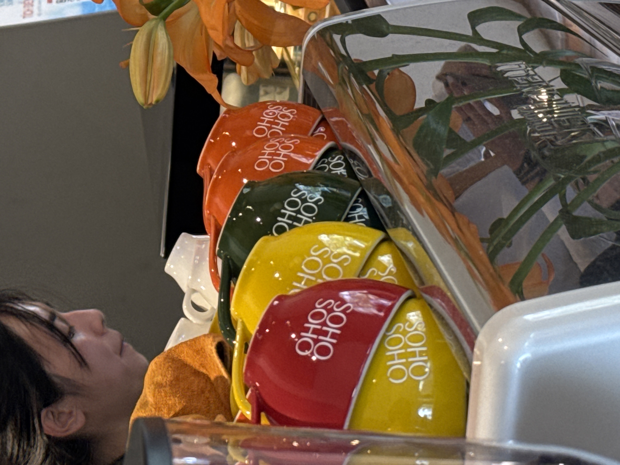 The image shows a close-up of several colorful mugs stacked on a counter. The mugs are red, yellow, and green, and each has "OHOS SOHO" printed in white lettering. In the background, a person's face is partially visible, with dark hair and a profile view. To the right, an orange flower is in bloom. The scene appears to be inside a cafe or shop, possibly a coffee shop, given the presence of mugs and the setting. There are no identifiable landmarks that place the scene in Da Lat, Vietnam; the description is purely based on the visual elements within the image.