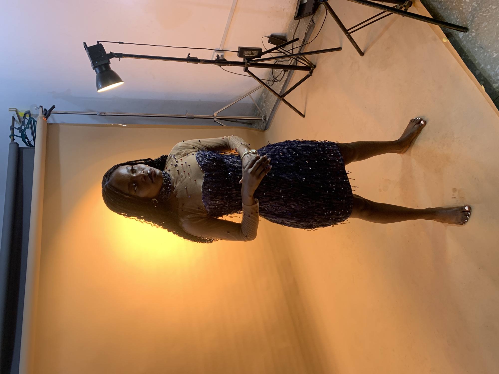 A full-length photograph depicts a Black woman in a short, dark, beaded dress with sheer long sleeves and a nude-illusion neckline, lying on her back. She is positioned on a backdrop that is a gradient of orange to beige, resembling a seamless paper roll studio setup. Her braided hair is spread out around her head. She is looking directly at the camera with her hands clasped near her chest.
In the background, studio lighting equipment is visible, including a large black lamp on a boom arm extending from a tall stand and another tripod stand holding what appears to be a power adapter. Other equipment is partially visible in the upper left corner.
The setting seems to be an indoor studio, and the lighting suggests a controlled environment for a professional photoshoot. There's no indication in the photo that the location is Gura Top, Nigeria, or any other specific place. The setting is clearly an indoor photographic studio.