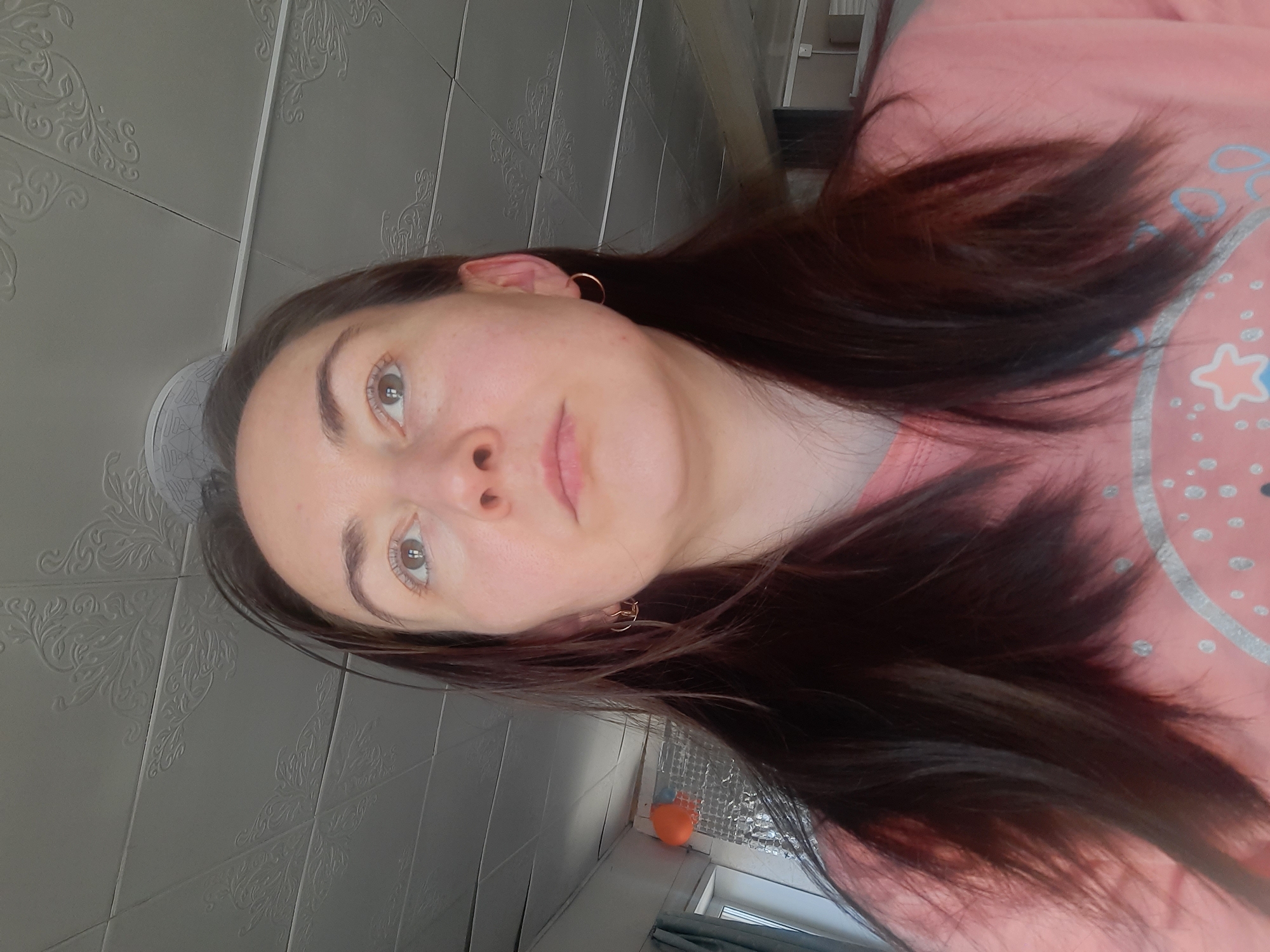 A woman with dark hair and fair skin is facing the camera. She is wearing gold hoop earrings and a pink shirt. The background includes a patterned wall, a window with curtains, and what appears to be an orange object on the windowsill. The location cannot be determined to be Krasnogorskoye, Russia from the provided image.