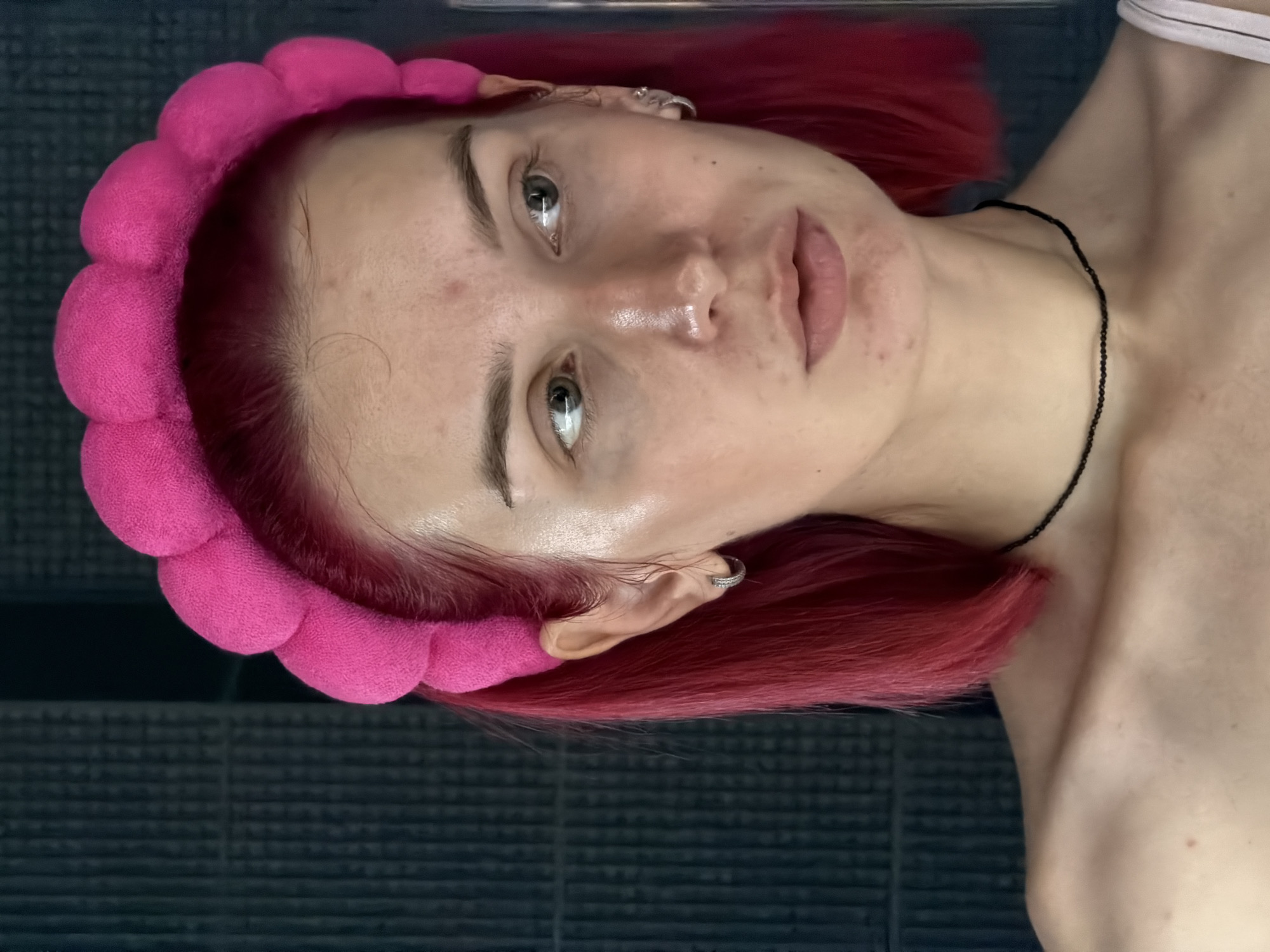 A young adult female with short, bright fuchsia-red hair and a wide, puffy, hot pink headband is depicted in a close-up, chest-up portrait. Her fair skin shows visible natural texture, light blemishes on her forehead, cheeks, and chin, and subtle dark circles under her greenish-hazel eyes. She wears small, silver-toned hoop earrings in both earlobes and a thin black beaded choker necklace. Her bare shoulders and prominent clavicle are visible, with a white clothing strap partially seen on her right shoulder. Her lips are slightly parted, and she looks towards her left with a neutral, contemplative expression. The background consists of a dark, uniform, textured surface with vertical grooves, suggesting an indoor setting. The scene is identified as being located in the city of Chaikovsky, Russia.