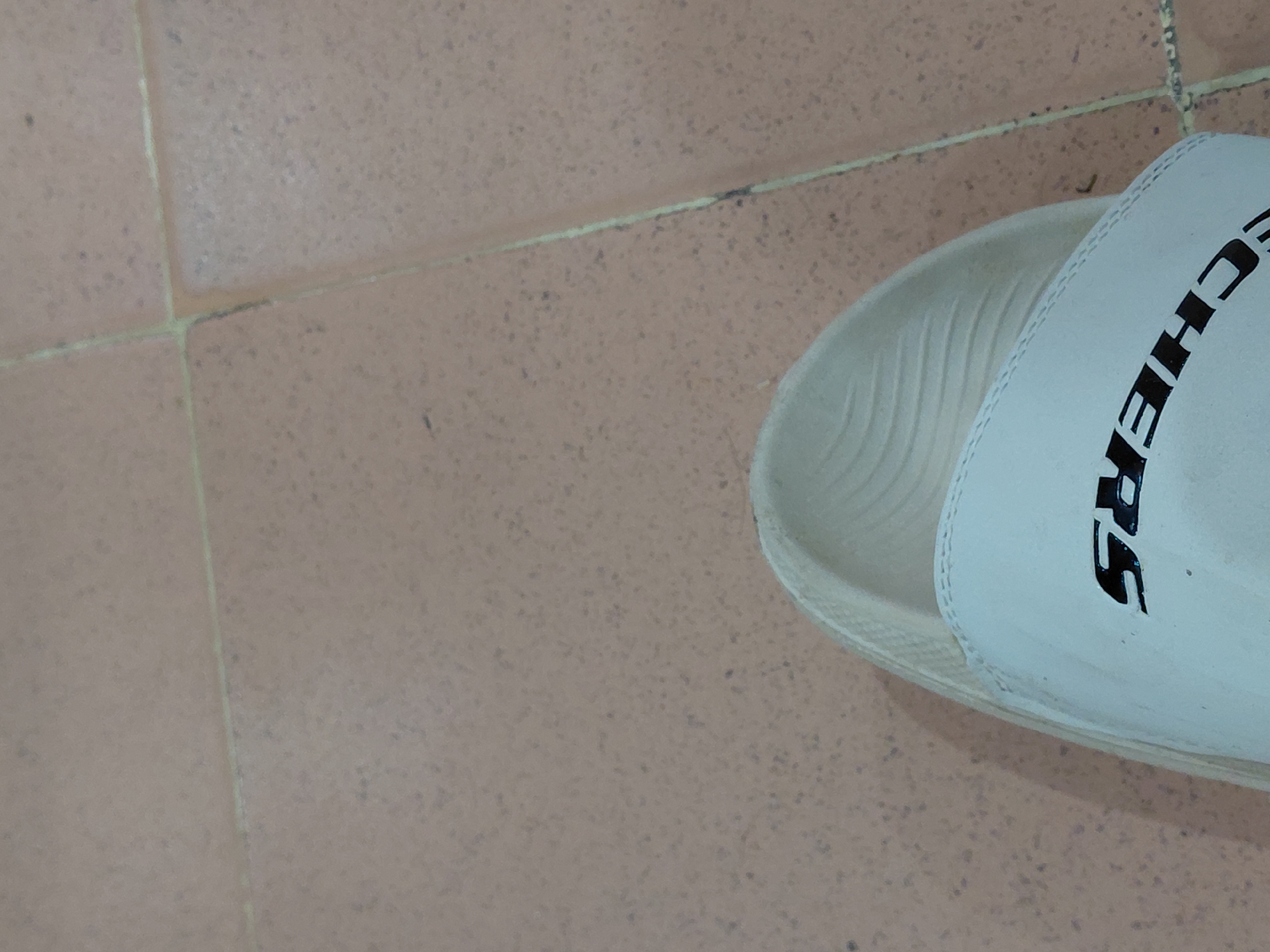 The image depicts a single, white Skechers slide on a tile floor. The tile appears to be a reddish-brown color with grout lines visible. The Skechers logo is partially visible on the upper strap of the slide. The photo is taken from an overhead perspective. There is no evidence to suggest this image was taken in Santa Ana, Venezuela.
