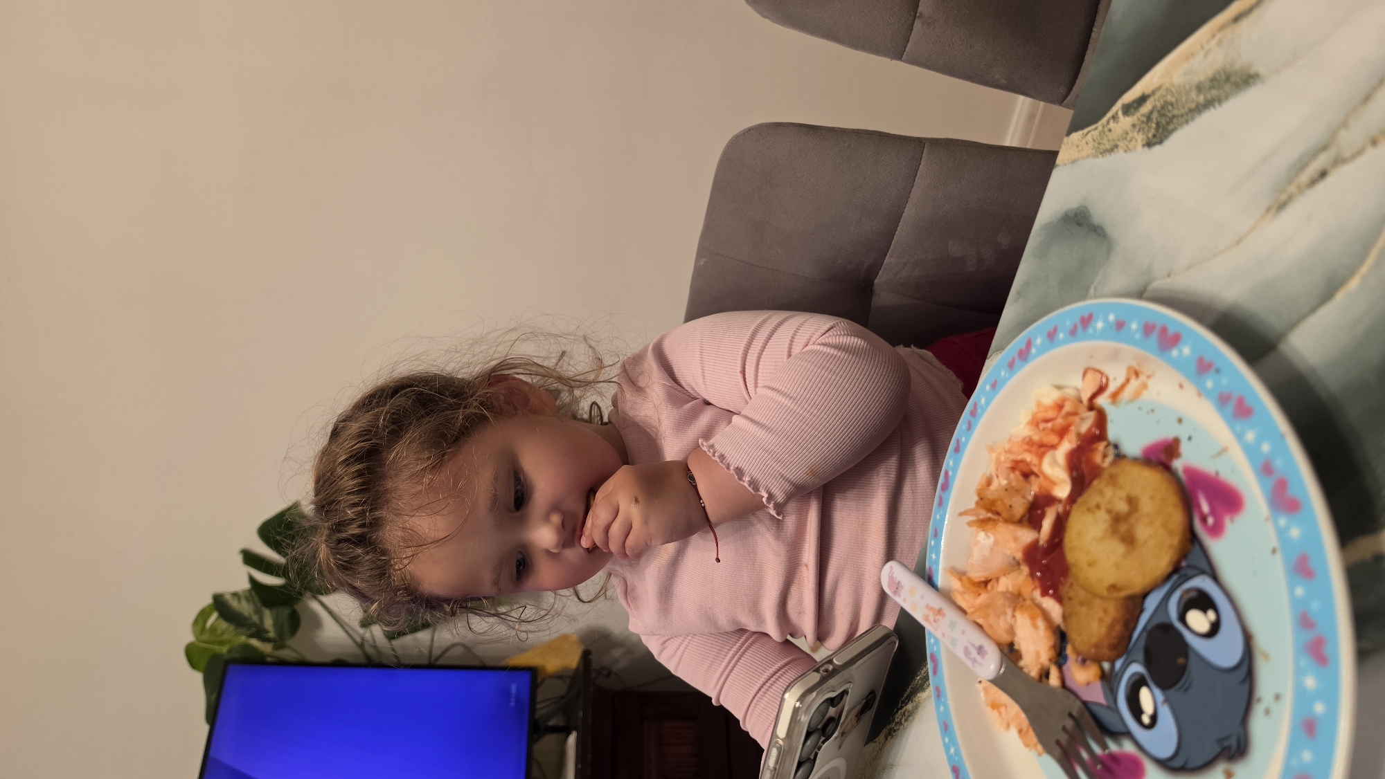 A young child with curly brown hair, wearing a long-sleeved pink shirt, sits at a table in an indoor setting. She is eating food from a blue plate with a Stitch character design. The plate contains what appears to be shredded chicken, hash browns, and a red sauce. Utensils are present on the plate. A smartphone is visible on the table next to the child. A gray upholstered chair is behind her. In the background, there is a television set and a plant. The location is not discernable from the image; therefore, it is impossible to determine if this image was taken in London, United Kingdom.