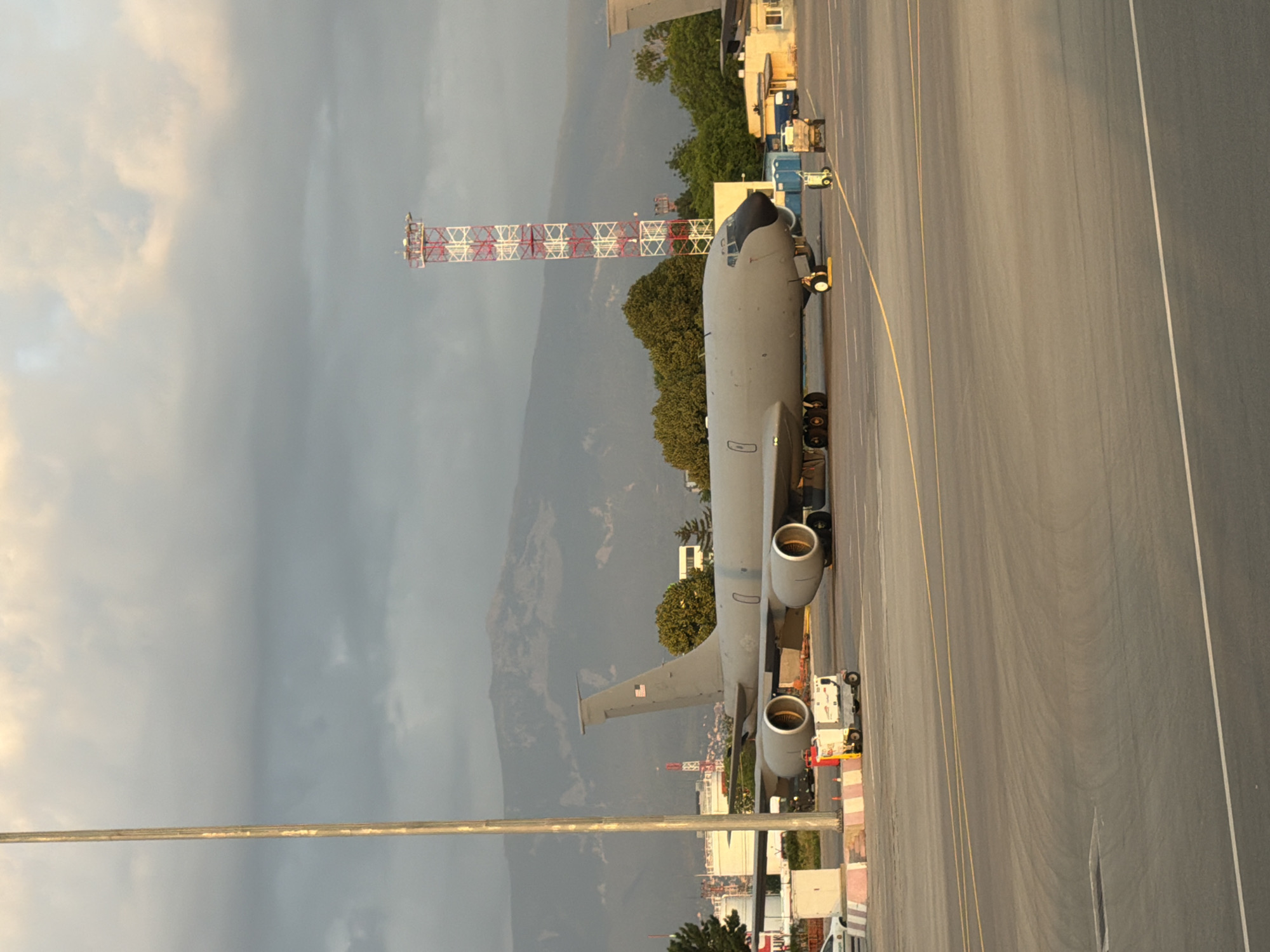 The image depicts a large, gray military cargo aircraft parked on an airport tarmac. The aircraft has four engines mounted under its wings and a distinctive nose. In the background, a towering red and white radio mast is visible, characteristic of the area around Sofia Airport. A mountain range looms in the distance, partially obscured by clouds. The scene is likely at Sofia Airport in Sofia, Bulgaria, given the visual cues. The tarmac is marked with yellow and white lines. Several pieces of ground equipment, including a small utility vehicle and a cart, are positioned near the aircraft. The sky is overcast with some lighter patches indicating the sun may be breaking through. The overall impression is one of a static scene at an operational airport.