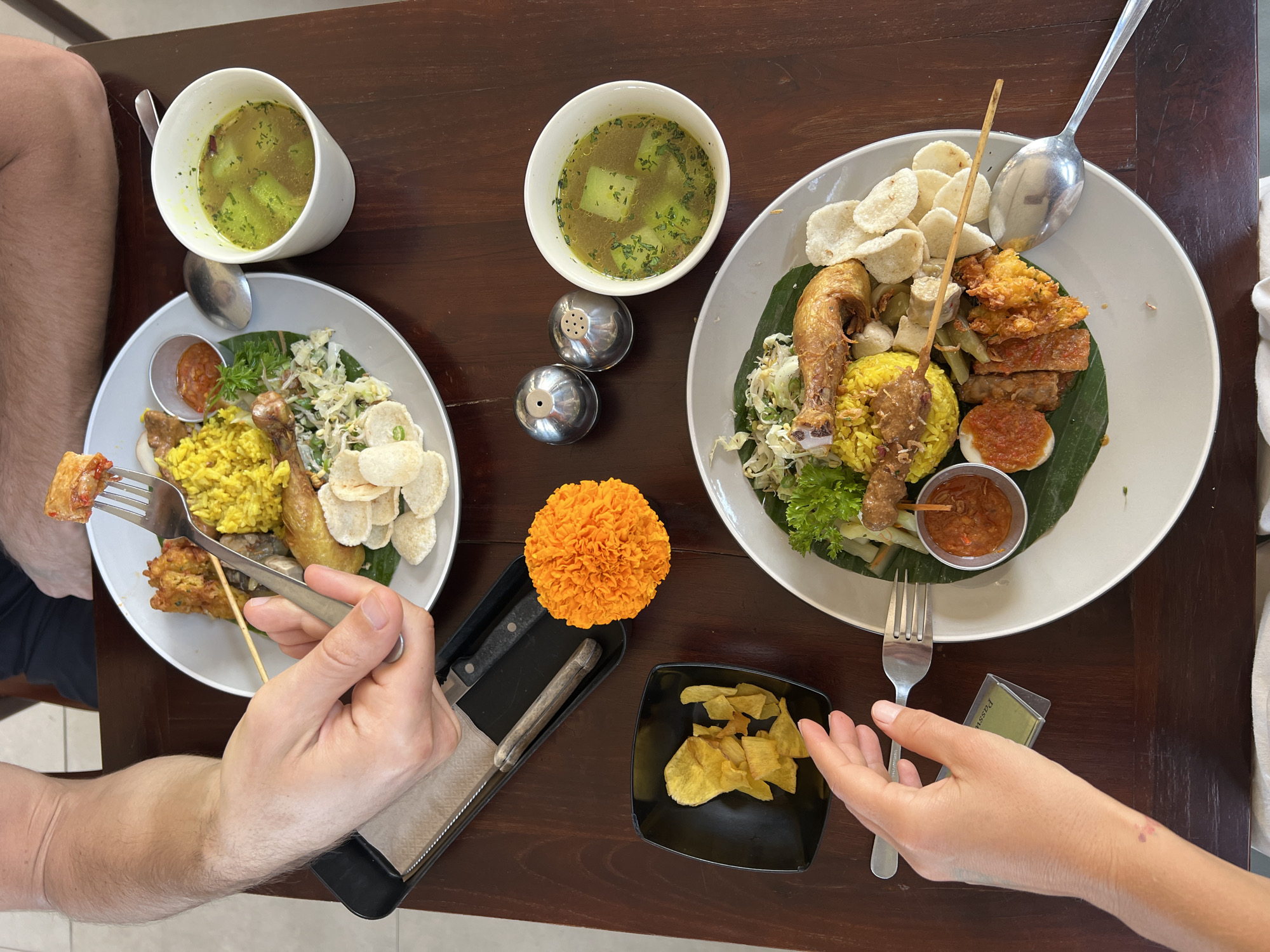 Two people are seated at a dark wooden table, eating a meal. The table is set with two white plates of food, two white cups of soup, a small black bowl of chips, a salt and pepper shaker set, and a decorative orange marigold. One person's arm is visible on the left side of the frame, holding a fork with food. The other person's arm is visible on the bottom left, reaching towards the bowl of chips. The plates of food appear to be a traditional Indonesian dish, likely Nasi Campur, consisting of yellow rice, fried chicken, vegetables, crackers, and a small dish of sambal. The setting appears to be a restaurant in Ubud District, Indonesia.