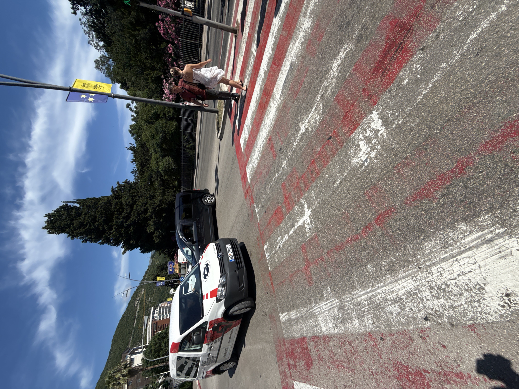 The image depicts a crosswalk with fresh red and white paint markings. Two people, a man and a woman, are walking across the crosswalk from right to left. They are dressed casually. A white van with red accents and the logo "ALFA GROUP TRANSPORT" is parked at the intersection, and a black van is behind it. In the background, there are trees, buildings, and mountains. The sky is blue with scattered clouds. This scene is located on a street in the city of Budva, Montenegro, likely near the coast given the presence of mountains and lush greenery.