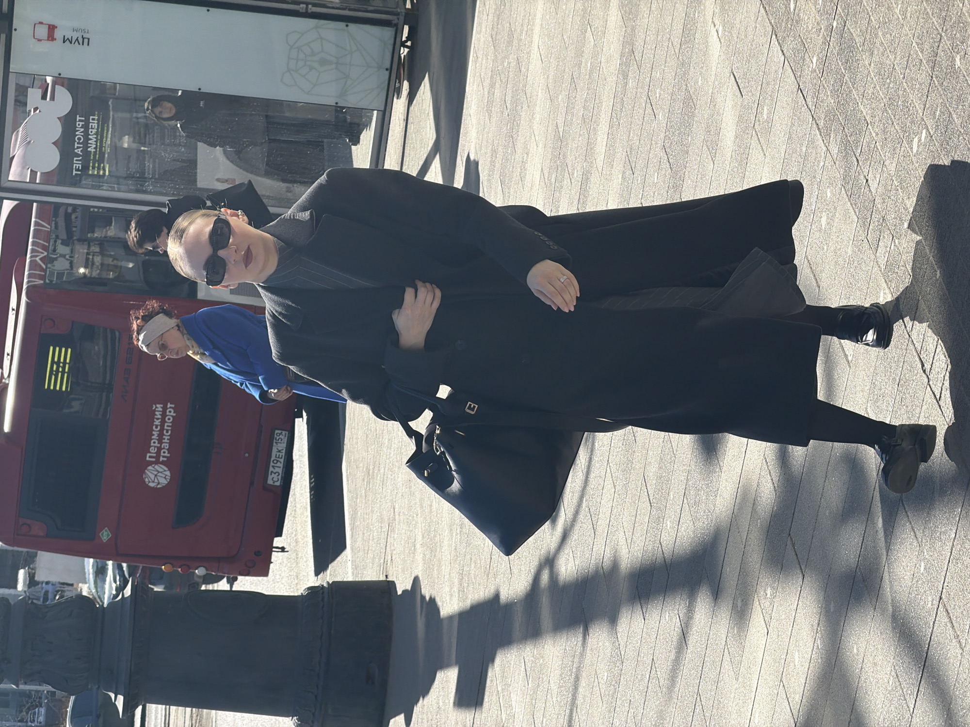 A person in a long black coat and sunglasses stands on a paved sidewalk in Perm, Russia, next to a red bus with Cyrillic text that translates to "Perm Transport," as well as a license plate. The person carries a black purse. Another person is walking behind them. Other people are inside a building in the background. A stone pillar is in the foreground.