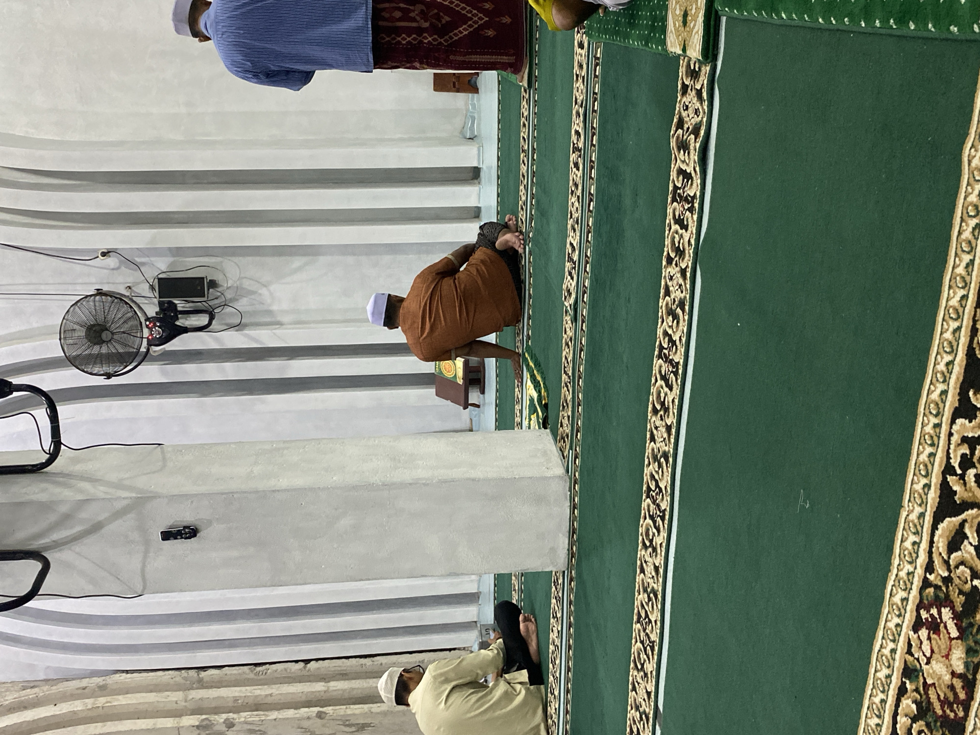 The image depicts the interior of a mosque in Lhokseumawe, Indonesia, characterized by its green carpeted floor with patterned borders. Three men are visible, dressed in traditional attire. One man, wearing a light-colored shirt and a white cap, is seated on the floor on the left side of the frame. Another man, in an orange shirt and patterned pants, is also seated on the floor in the middle of the image, facing away from the camera. A third man, wearing a blue striped shirt and a sarong, stands facing away from the camera on the right side of the frame. The mosque's architecture features tall, fluted pillars and a white, striped wall. A ceiling fan is mounted on the wall, along with what appears to be a remote control attached to the wall with adhesive. A small wooden stand with a book is positioned behind the seated man in the orange shirt. The overall impression is one of a serene and devout atmosphere.