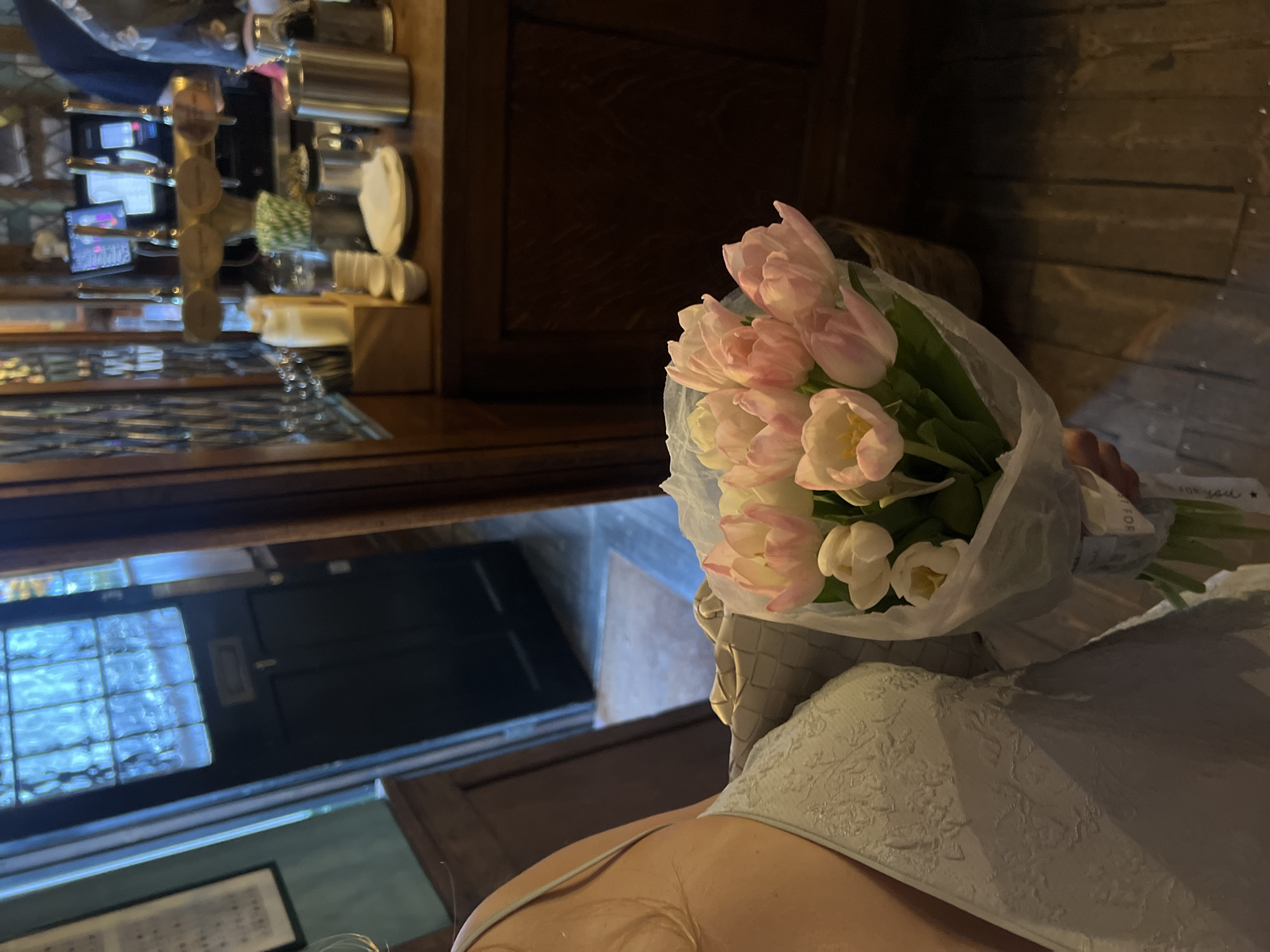 The image shows a person holding a bouquet of pink and white tulips wrapped in white paper. The person is wearing a light blue lacy top. In the background, there is a blurred view of what appears to be a bar or restaurant area. A barista counter with coffee-making equipment is visible, and a dark wooden door and window can be seen further in the background. The environment suggests an interior location in London, United Kingdom, based on the architectural style and the style of the bar/restaurant.