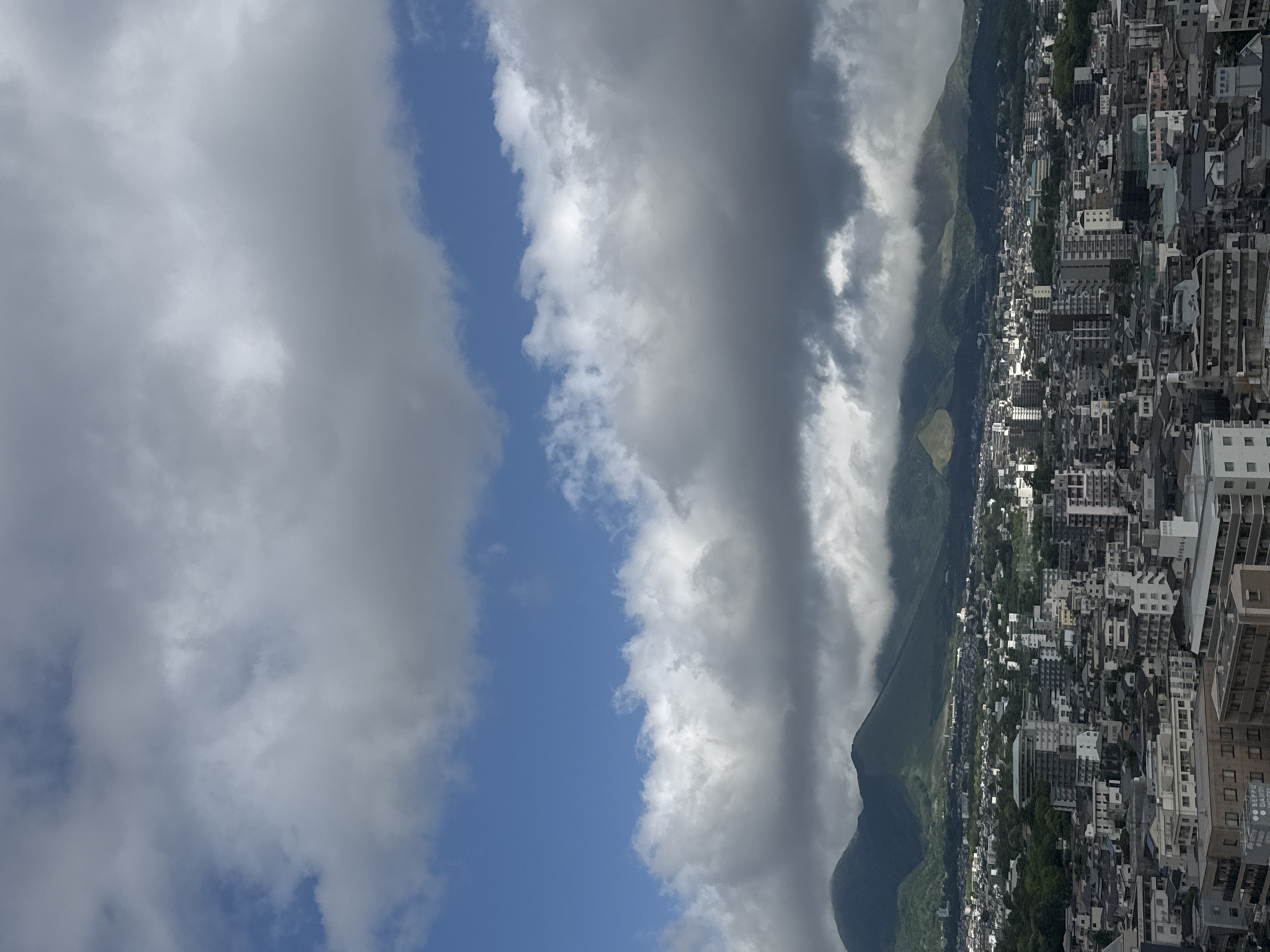 The image is an aerial view in Beppu, Japan, looking toward a cityscape densely packed with low to mid-rise buildings. A prominent mountain range rises to the left, partially obscured by heavy, dark clouds. The sky transitions to a mix of light blue and white clouds, with open patches of blue sky on the left side. The overall scene suggests a city near a mountainous region experiencing cloudy weather.