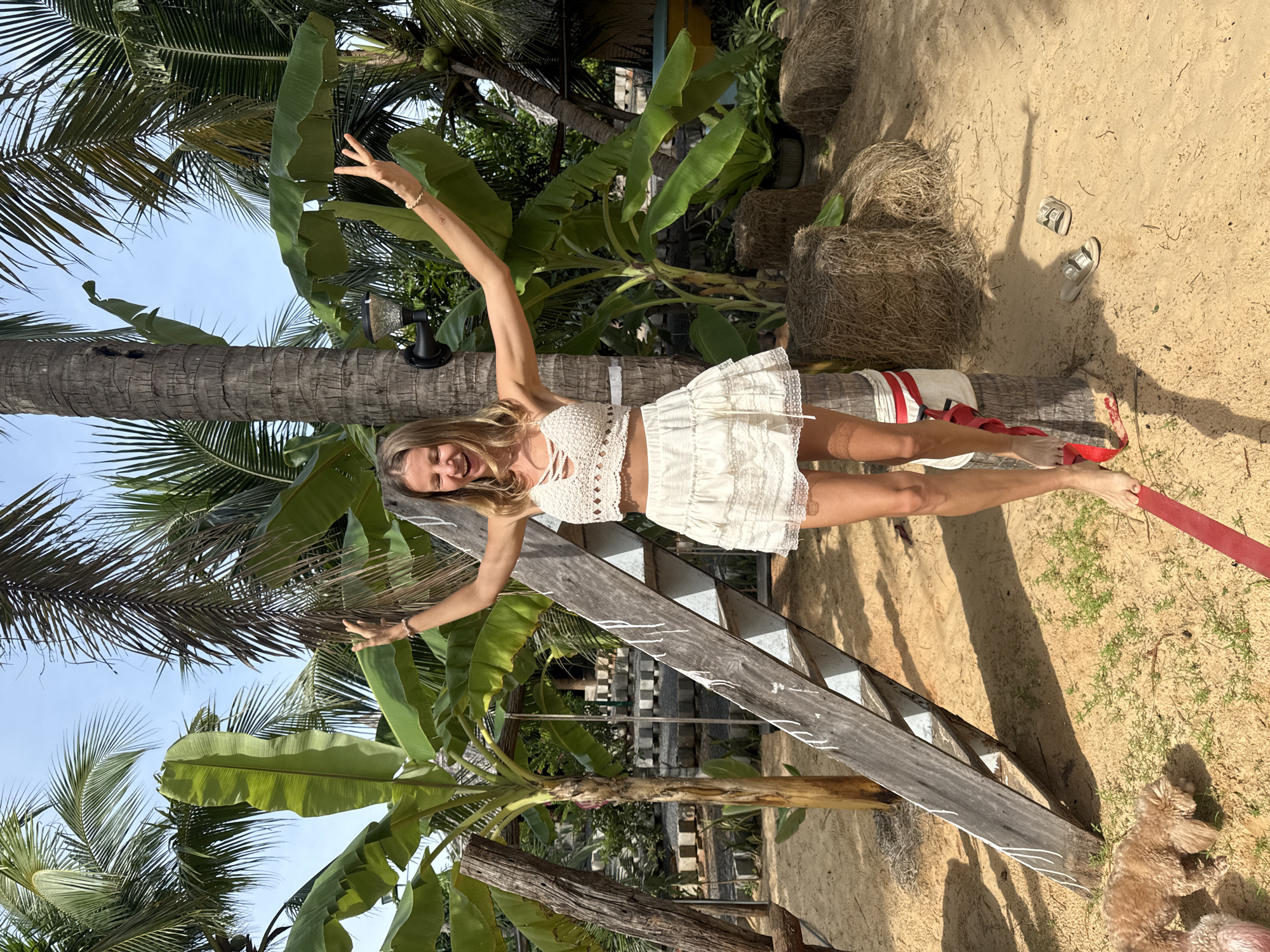 The image shows a young Caucasian woman with blonde hair, wearing a white crochet top and a white tiered skirt, balancing on a red slackline attached to a palm tree. Her arms are outstretched, and she is smiling. She is barefoot. In the background, there are lush green palm and banana trees, hay bales, and a rustic wooden ladder leaning against another palm tree. A small, light brown dog is visible in the lower left corner, sniffing the sand. A pair of light-colored sandals are on the sand to the right of the slackline. The scene appears to be a tropical beach setting, possibly in Mũi Né, Vietnam, given the vegetation and sandy ground. The lighting suggests it is a sunny day.