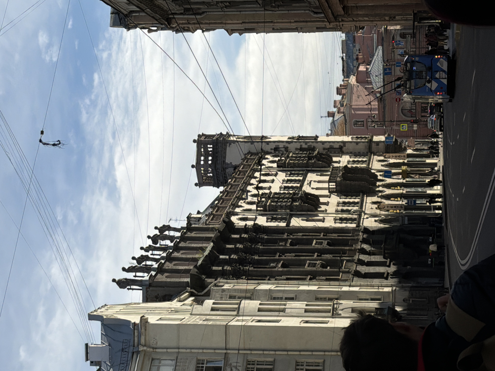 The image depicts a street scene in Saint Petersburg, Russia, notable for its architectural details. The camera is positioned looking upwards towards the buildings lining a street with a trolley bus visible on the right side. The building dominating the view has an ornate facade with statues and decorative elements along its roofline. Electrical wires crisscross the sky above. The sky is partially cloudy. A portion of a person's head and shoulder are visible in the bottom right corner, suggesting the photo was taken by someone standing on the street.