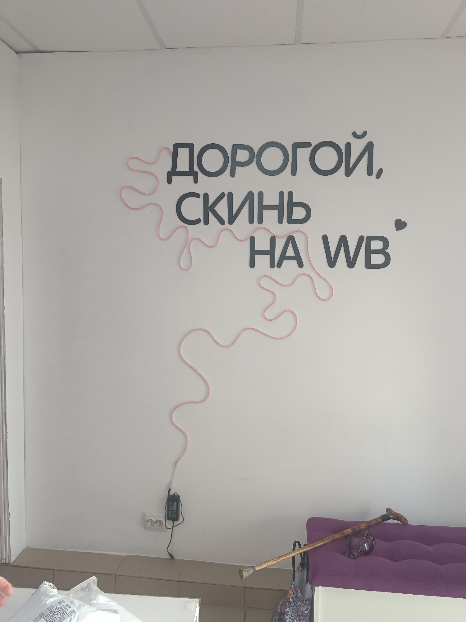 The image shows an interior view of a room with white walls and a tiled floor. On one wall, there are dark gray stylized letters spelling out "ДОРОГОЙ, СКИНИЬ НА WB" ("Dear, throw on WB" in Russian), decorated with a pink squiggly line that visually connects the words. Below the lettering, near the floor, there is an electrical outlet and a black adapter plugged in. To the right, there is a purple bench seat with a wooden cane leaning against it.