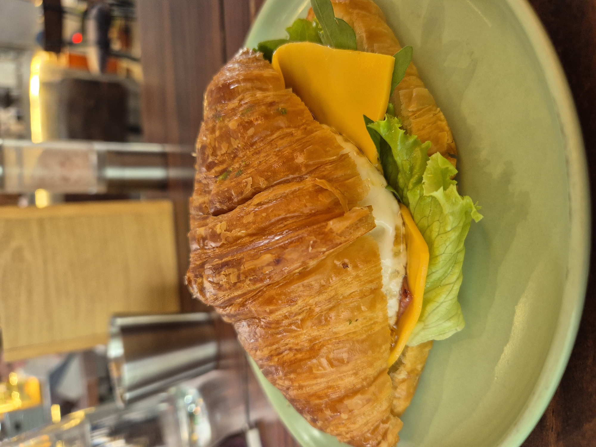 An overhead close-up shot shows a croissant sandwich on a light green plate. The croissant is golden brown and flaky, sliced in half to reveal the filling. The filling includes lettuce, a slice of yellow cheese, and what appears to be a fried egg. The sandwich is positioned slightly off-center to the right. In the background, out of focus, is a wooden table, a metal cup, a glass of water, and a wooden menu or sign. The overall setting suggests a cafe or restaurant environment. This scene is likely located in Đà Nẵng, Vietnam.