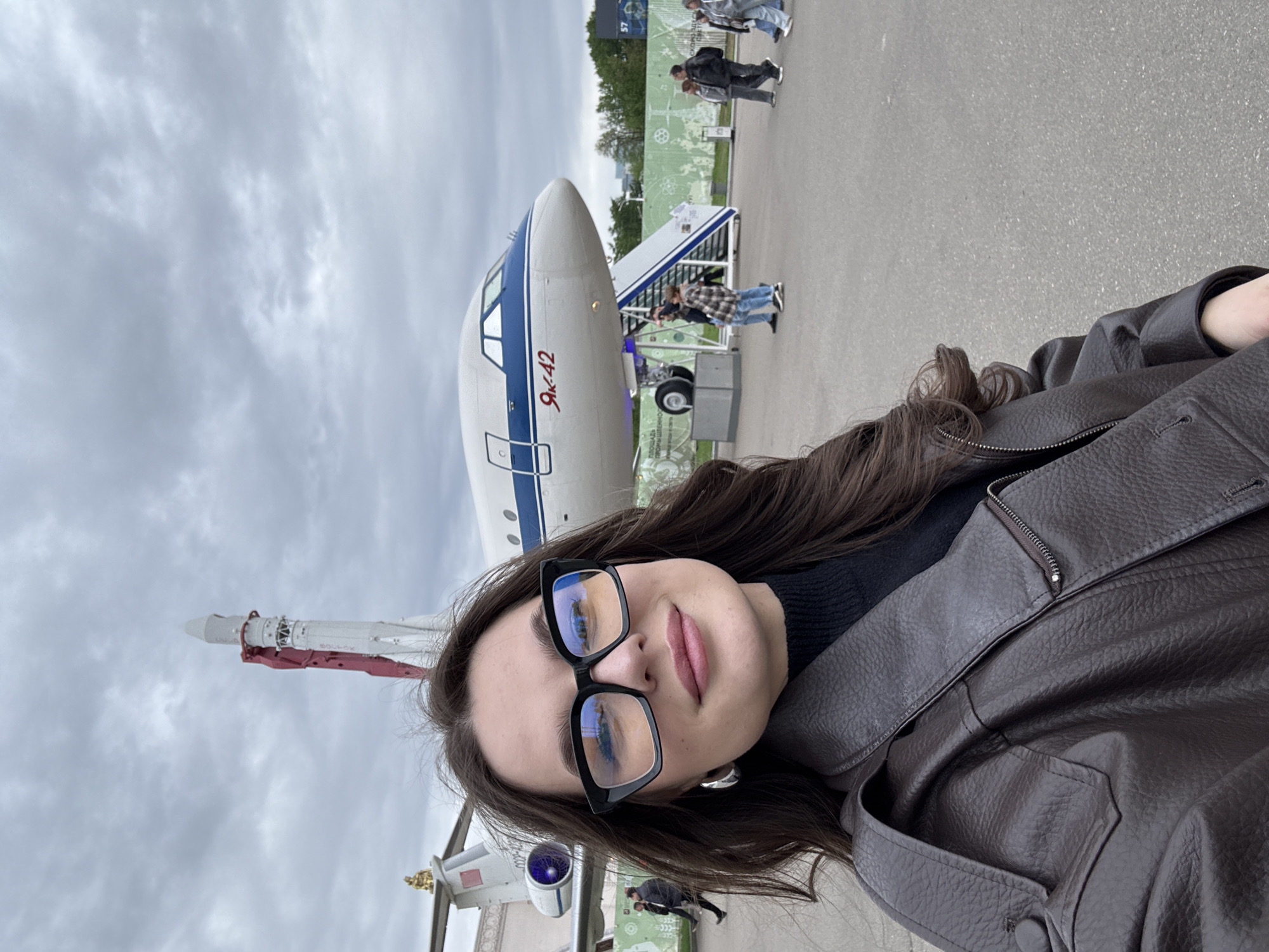 A woman is in the foreground, wearing glasses and a brown jacket. Behind her is an aircraft with the Cyrillic letters "Як-42" on its side. Behind the aircraft, there are several people. Further in the background, there is a tall rocket. The location is Moscow, Russia.
