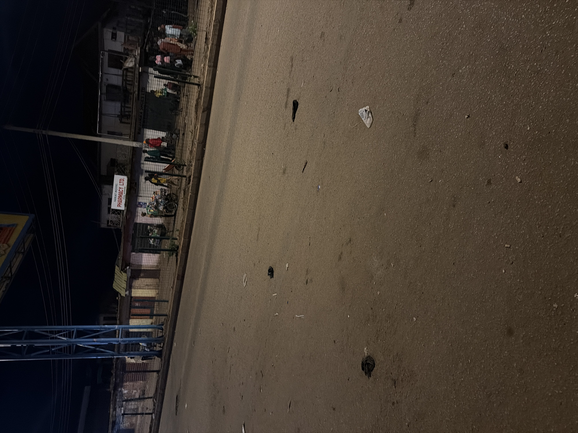 The image depicts a street scene at night in Bukuru, Nigeria. The foreground is dominated by a paved road with scattered debris, including what appears to be plastic bags and paper. A sidewalk runs along the left side of the road, lined with a metal fence. Beyond the fence are several small shops or stalls, some with corrugated metal roofs. One shop has a sign that reads "MERAT MODEL PHARMACY LTD." In the background, a few people are visible, some standing near the shops and others walking. A motorcycle is also present, and a cart with wheels is near the pharmacy. The overall environment suggests a commercial area at night, with limited lighting and evidence of litter on the road.