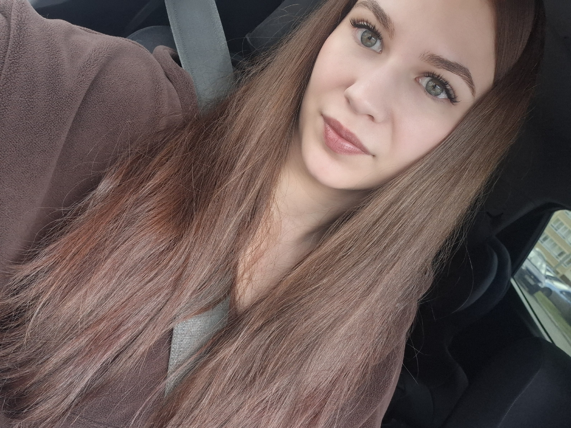A young woman is captured in a selfie-style close-up, seated inside a car in Chaikovsky, Russia. She has long, straight, brown hair with noticeable reddish-auburn undertones, which cascades over her left shoulder and chest, framing her face. Her light green or hazel eyes are prominent, enhanced by long, dark eyelashes and well-defined brown eyebrows. Her skin appears fair and smooth, and her full lips are tinted with a light pinkish-brown lipstick or gloss. She wears a dark brown top made of a textured, possibly fleece-like fabric.
The woman looks directly at the camera with a calm, subtle smile. A light grey seatbelt crosses over her left shoulder, indicating she is in the passenger or driver's seat of a vehicle. The car's interior appears dark, with glimpses of dark upholstery. Through the window on the right side of the image, residential buildings, likely apartment blocks, are partially visible, along with other parked cars, suggesting an urban or suburban street scene during daytime. No text is visible in the image.