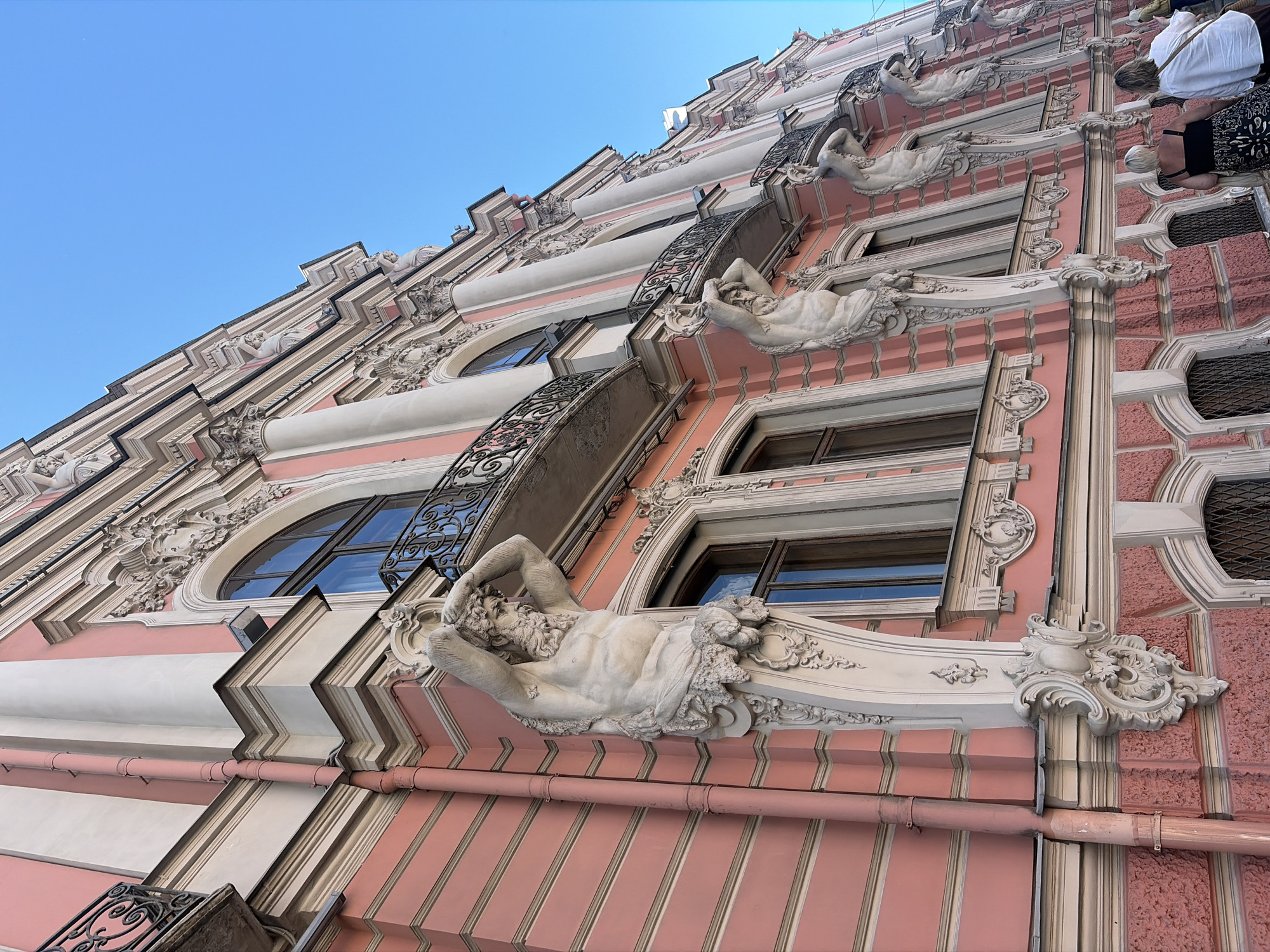 The image shows the facade of a pink and cream-colored building in Saint Petersburg, Russia, with ornate architectural details. The building features multiple stories with arched windows, balconies with decorative wrought-iron railings, and classical columns. Prominently featured are several life-sized sculptures of muscular male figures supporting the upper portions of the building, specifically the balconies. These figures, likely caryatids or atlantes, are depicted in a strenuous pose with their arms raised. The style of architecture suggests a Baroque or Neoclassical influence. The sky above is clear blue. In the foreground, two women are walking away from the camera, their backs to the viewer, suggesting they are tourists or passersby. The overall impression is of a grand and historic urban setting.
