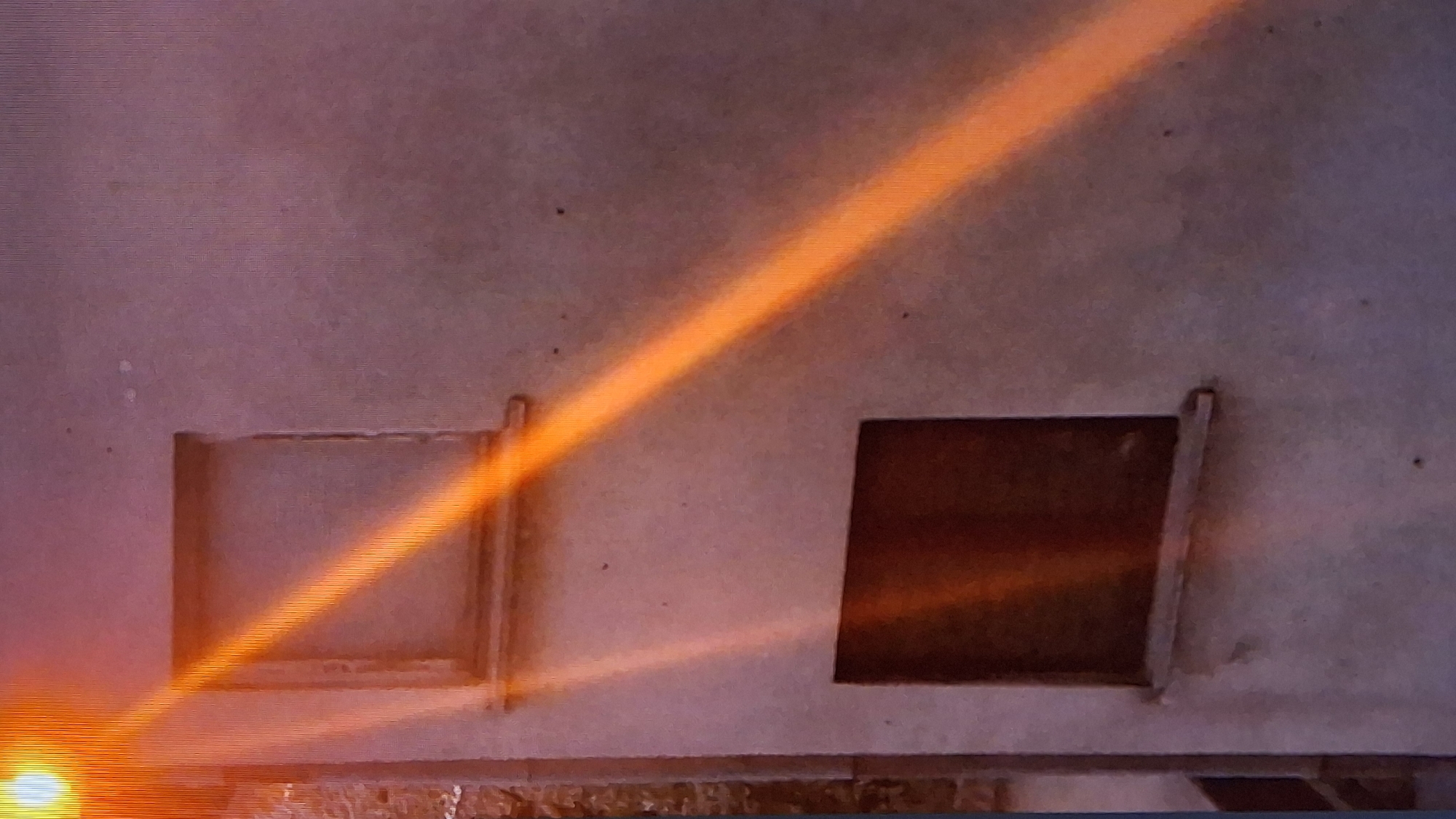 Here is a forensic description of the image:
**Scene:** Indoor setting with two square openings in a wall or structure. One opening appears brighter than the other.
**Object:** An orange, light-like beam diagonally illuminates the image.
**Environment:** The wall is textured, with some specks and color variations. The light source casts intense illumination, particularly on the lower-left side.