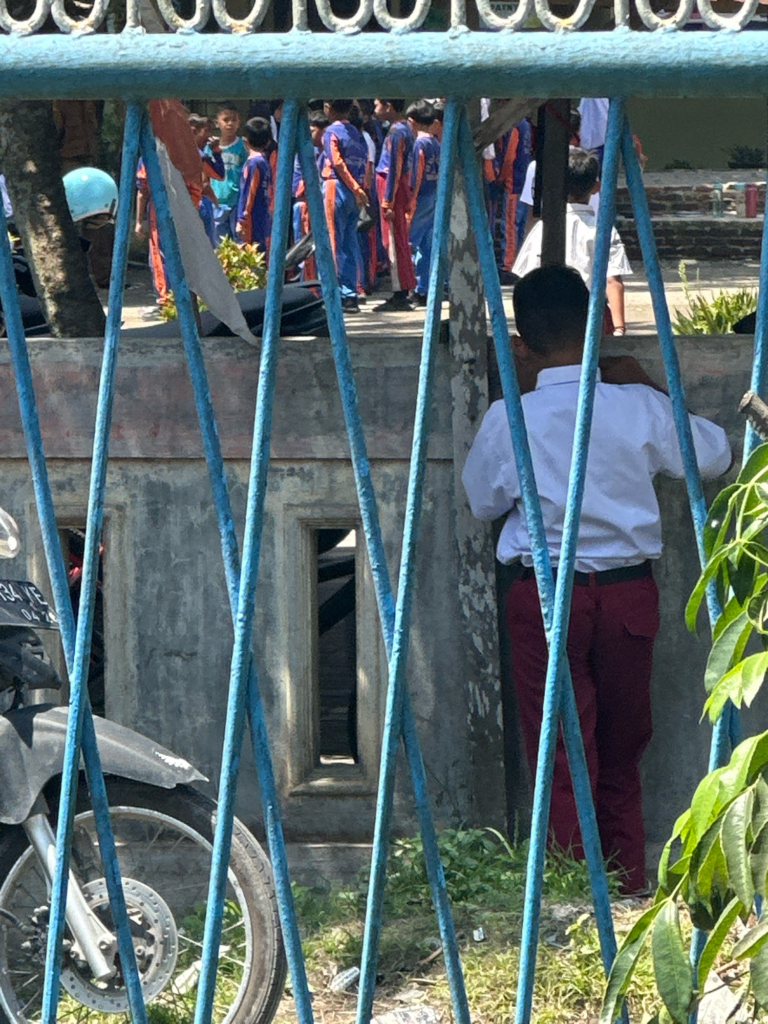 The image depicts a young boy in a white shirt and maroon pants standing with his back to the camera, looking towards a group of children in similar school uniforms. He is positioned in front of a weathered, gray concrete wall with a narrow, rectangular opening. The scene is viewed through a blue metal fence. To the left, a portion of a parked motorcycle is visible. In the background, several children are standing in what appears to be a schoolyard or public area, some wearing blue and orange uniforms. The ground is covered with grass and debris. The overall impression is of a school environment in an outdoor setting. The location is identified as the City of Binjai, Indonesia.