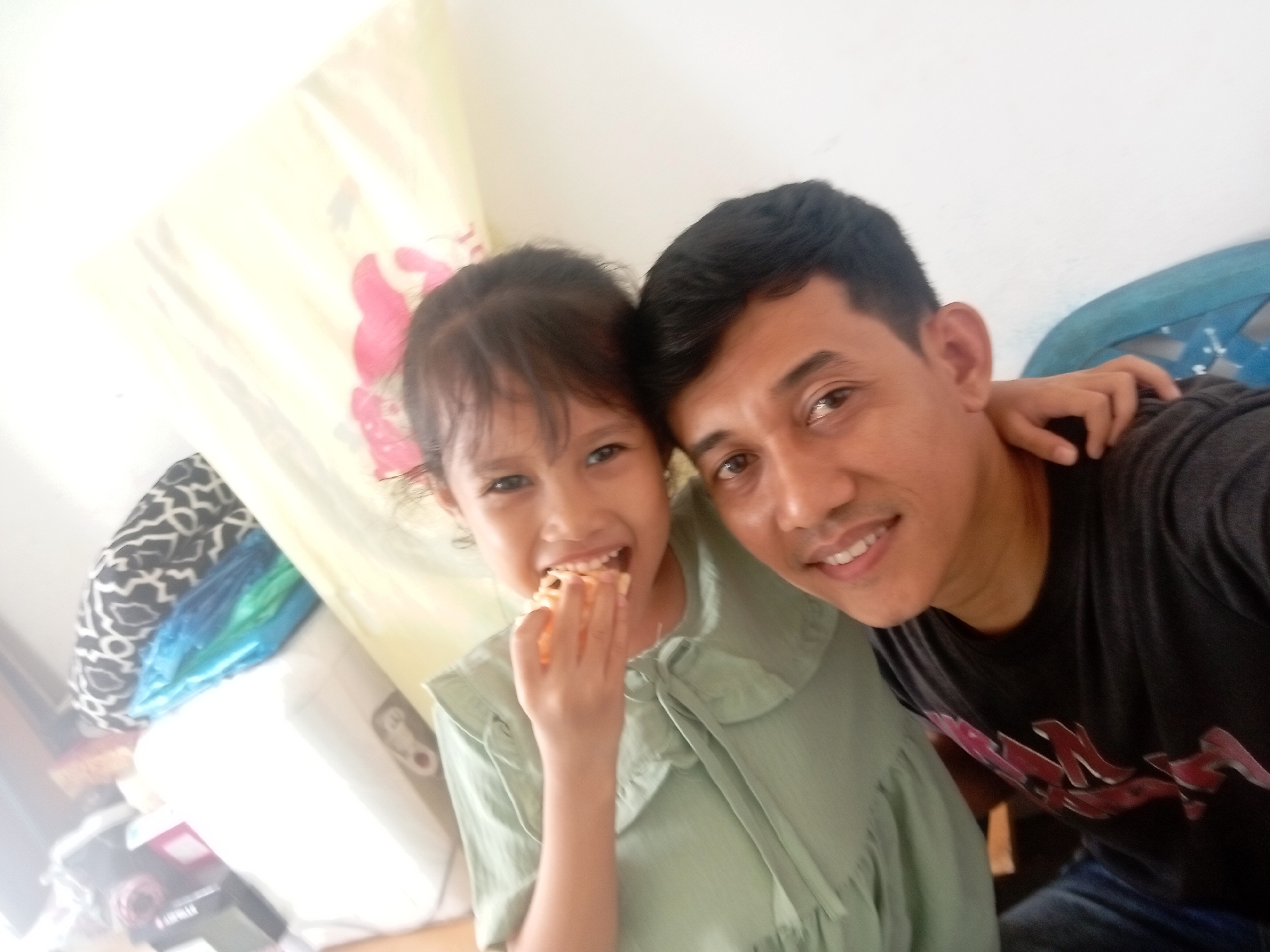 This is a selfie of a father and his young daughter. The father, on the right, has dark hair and is wearing a dark t-shirt with a red graphic design. He is smiling at the camera. The daughter, on the left, has dark hair and is wearing a light green dress with a ruffled collar. She is eating a piece of food, possibly a cracker or cookie, and smiling. Her left arm is around her father's neck.
The background is blurry but appears to be an indoor setting. There is a light-colored curtain or fabric with a floral pattern behind the daughter. To the left, there are some stacked objects, including a white cylindrical item and a patterned textile. To the right, a blue object, possibly a chair or piece of furniture, is visible.
The lighting suggests it is daytime. The overall mood is cheerful and affectionate, capturing a candid moment between father and daughter. There is no visible text in the image. The location context is Batam, Indonesia.