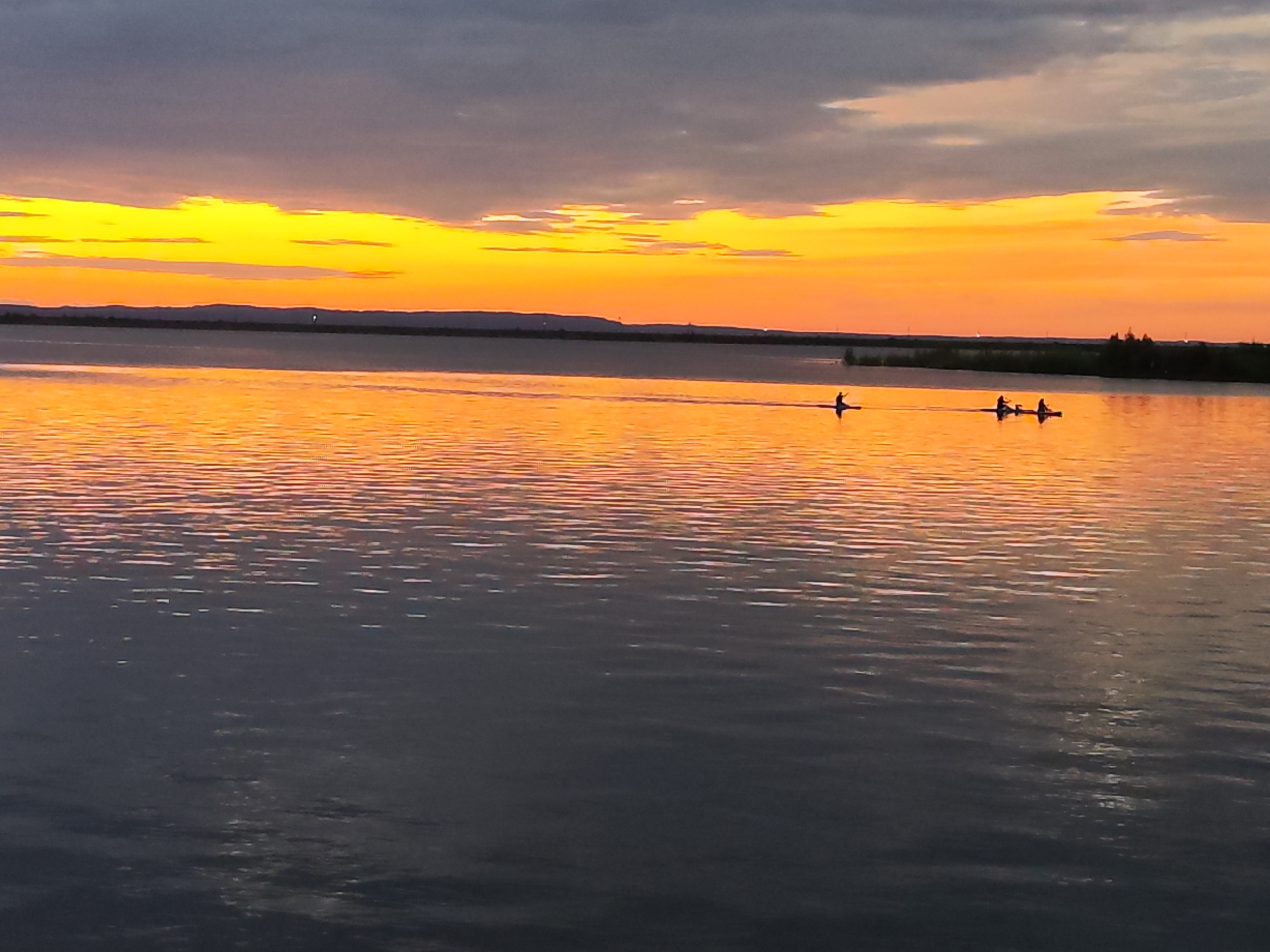 This is a horizontal, medium-shot photograph taken at sunset over a body of water, likely a river or lake, near Chaikovsky, Russia. The sky is a dramatic blend of dark, cloudy gray at the top, transitioning to vibrant bands of bright orange and yellow closer to the horizon. The setting sun casts a warm, golden-orange glow across the water, creating shimmering reflections.
The scene features three individuals in kayaks, spaced apart and paddling across the calm water. One kayaker is in the left foreground, while two others are further to the right and closer together. They appear to be enjoying a recreational activity as the day ends. The distant shoreline is visible as a dark, silhouetted line with faint hints of trees and possibly some structures.
The overall mood of the image is serene and peaceful, amplified by the warm colors of the sunset and the quiet activity of the kayakers. The time of day is clearly sunset, and the weather appears to be fair with dramatic cloud formations. There is no visible text in the image.