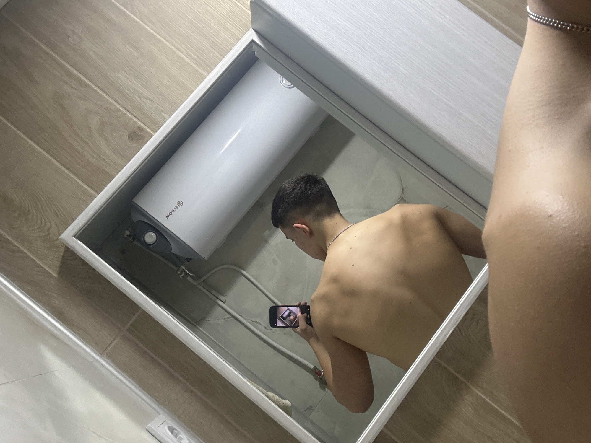 A photograph taken in an interior, possibly a bathroom, in Zlatograd, Bulgaria. The image is a reflection in a mirror of a shirtless, light-skinned man holding a smartphone. The man appears to be taking a photo of himself in the mirror. In the background, behind the man's reflection, is a cylindrical, white water heater mounted on the wall. The walls and floor appear to be tiled in light brown and beige. The location in the city of Zlatograd cannot be independently determined from this image alone.