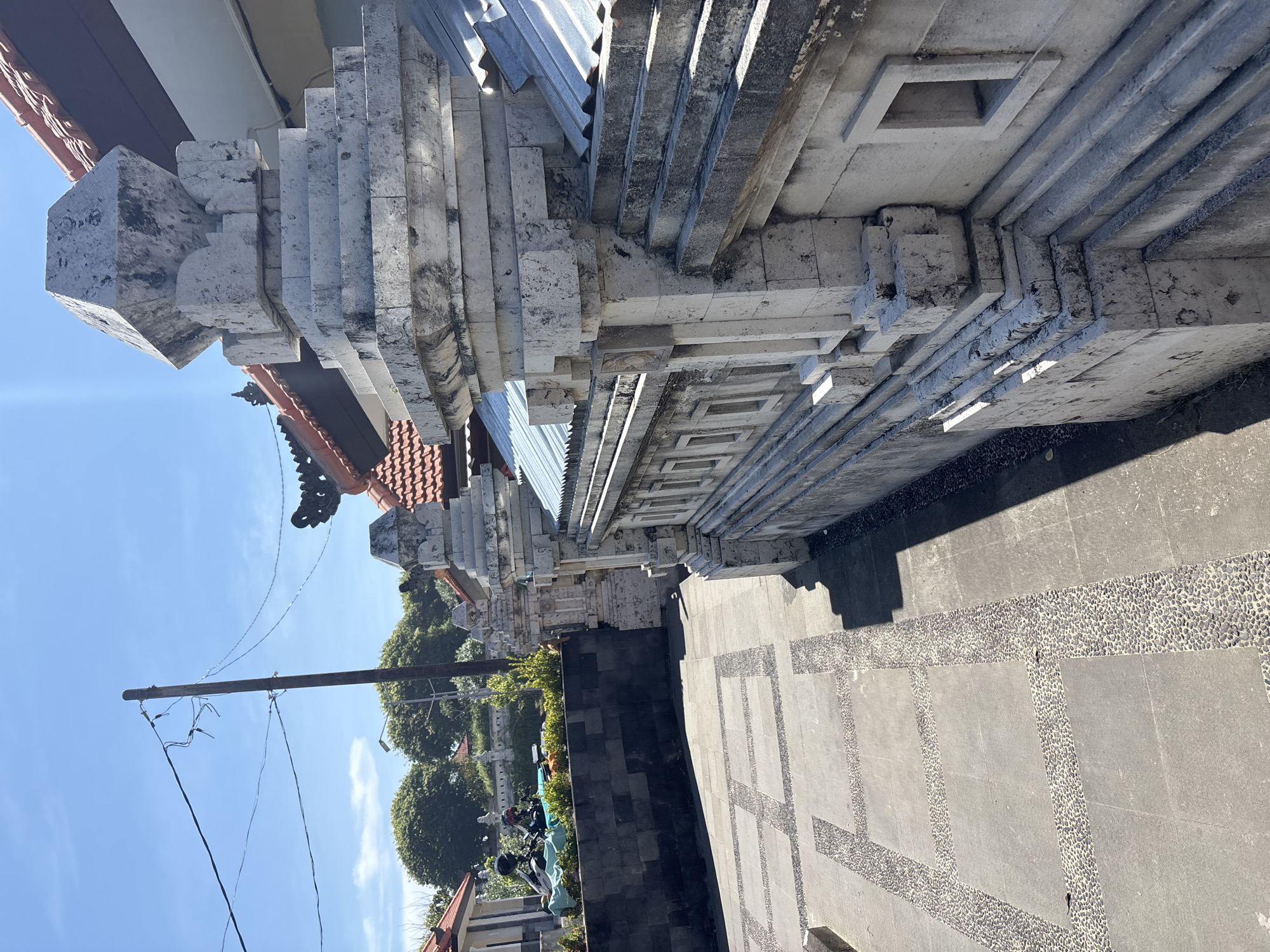 The image depicts a building in Kuta Selatan, Indonesia, characterized by tiered, white stone architecture. The building's façade features layered horizontal elements and square decorative recesses. A tiled roof is partially visible. The ground is paved with grey stone tiles interspersed with decorative pebble strips. A low black wall separates a landscaped area with greenery and parked motorbikes. In the background, trees and buildings are visible under a clear blue sky. A utility pole with wires is prominent in the middle ground.