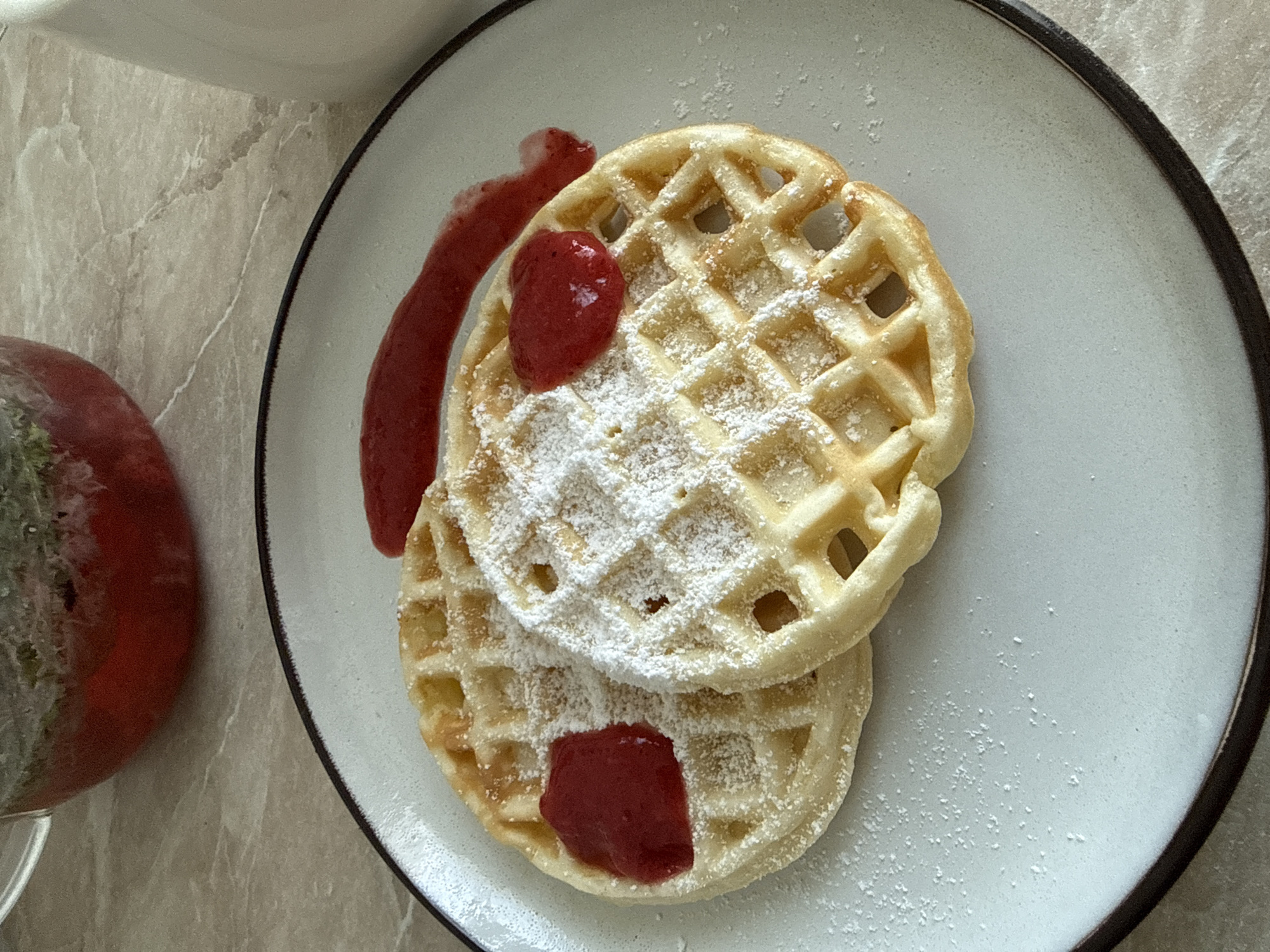 A close-up shot depicts a stack of two waffles on a light blue plate with a dark rim. The waffles are dusted with powdered sugar and topped with red fruit sauce or compote. A portion of the sauce is arranged as a smear on the plate adjacent to the waffles, with another dollop on the top waffle and a similar dollop on the bottom waffle.
To the left, a clear glass teapot filled with a red beverage is partially visible. The surface beneath the plate appears to be a light-colored stone or imitation stone countertop. The exact location in the city of Chaikovsky, Russia, cannot be determined from the given image.