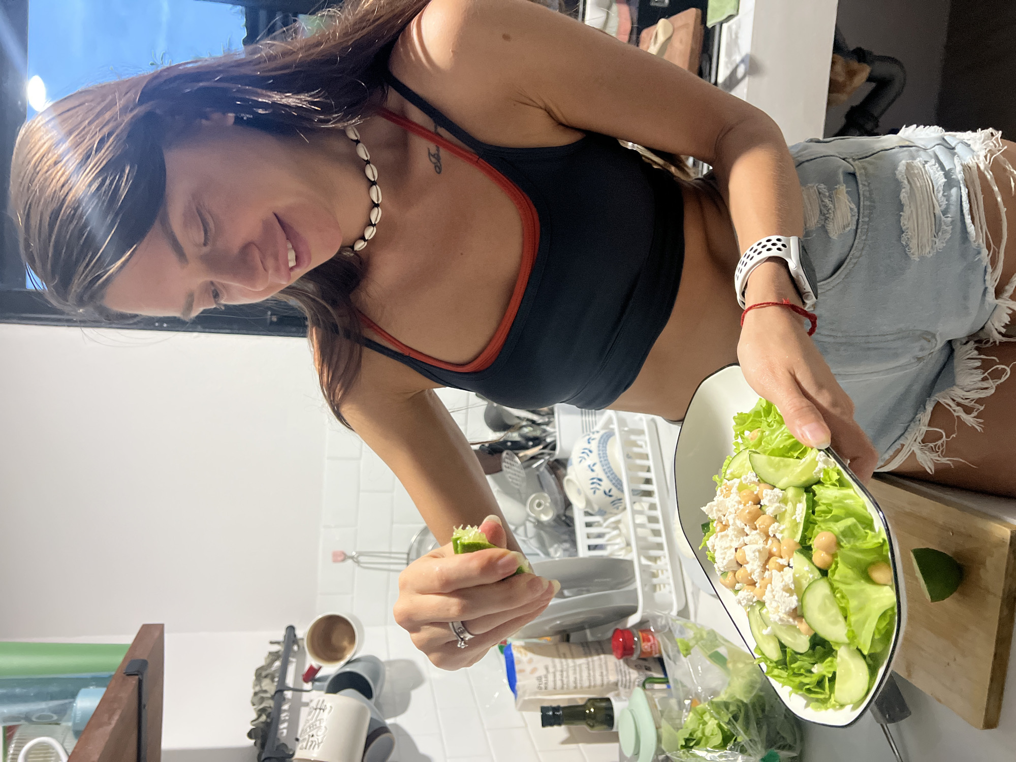 A woman is preparing a salad in a kitchen. She is wearing a black and orange sports bra, ripped denim shorts, a shell necklace, and a white smartwatch. She is squeezing a lime into a white bowl filled with lettuce, cucumbers, chickpeas, and white cheese. The kitchen background shows a white tiled wall, a dish rack filled with dishes, and various food items. The location is potentially บ้านบางรักษ์, Thailand, though there is no definitive evidence within the image.