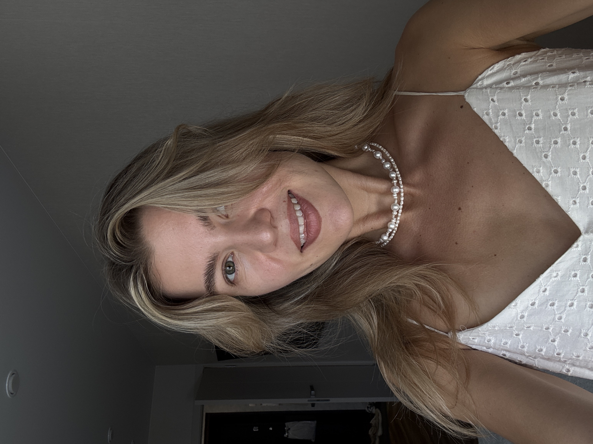 A woman with blonde hair and green eyes is shown in a close-up shot, wearing a white broderie anglaise top and a pearl necklace. She is smiling slightly and looking towards the camera. The background is indistinct, with a wall and a doorway visible. The lighting suggests an indoor setting.