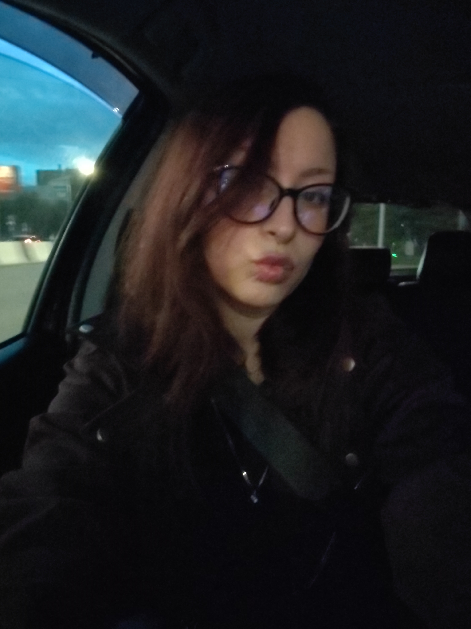 In this selfie taken inside a car, a young woman with auburn hair and glasses is looking at the camera with her lips pursed. The interior of the car is dark, but the blue hue of the sky is visible through the window, suggesting it is either dusk or dawn. The woman is wearing a dark jacket. The background outside the car shows glimpses of city lights and buildings, hinting at an urban environment, possibly Krasnoyarsk, Russia, given the context. The image is dimly lit, with the main focus on the woman's face.