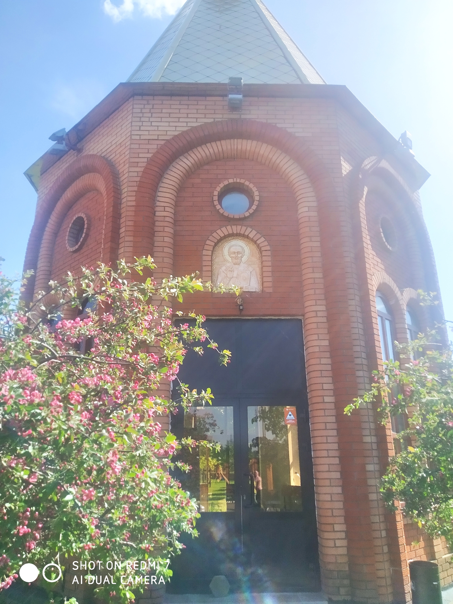 The image depicts a small, red brick building with a green, conical roof, located in Chaikovsky, Russia. The building's architecture features arched windows and a central entrance with double doors. Above the entrance, there is a circular window and a religious icon. The entrance is flanked by flowering trees with pink blossoms. The image was taken on a sunny day, as indicated by the bright sky. The lens of the camera partially obscures the scene. Text at the bottom indicates the image was taken with a Redmi 7 AI Dual Camera.