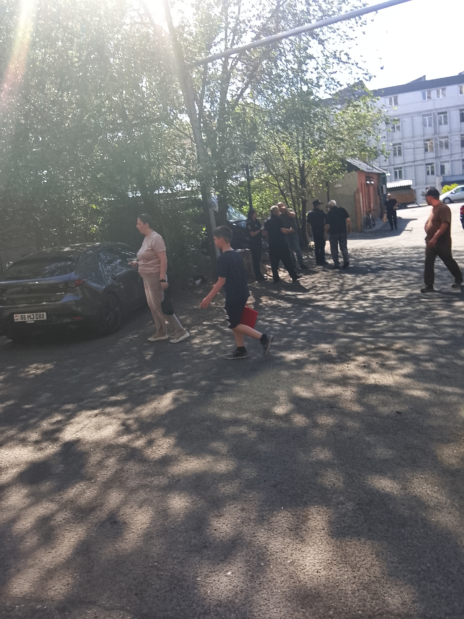 In a sun-drenched outdoor scene in an urban setting, a group of individuals is gathered. A dark-colored car is parked to the left, with a license plate reading "88 MJ 088". A woman, wearing light-colored pants and a light-colored top, stands next to the car, facing left. To her right, a young boy in a dark t-shirt and shorts walks away from the car, carrying a red object in his right hand. Behind the car and the woman, a group of men, mostly dressed in dark clothing, are standing and conversing. Further in the background, a multi-story building with numerous windows is visible, as well as a smaller structure with a red door. The ground is paved with asphalt and is partially covered in dappled shadows from overhead trees.