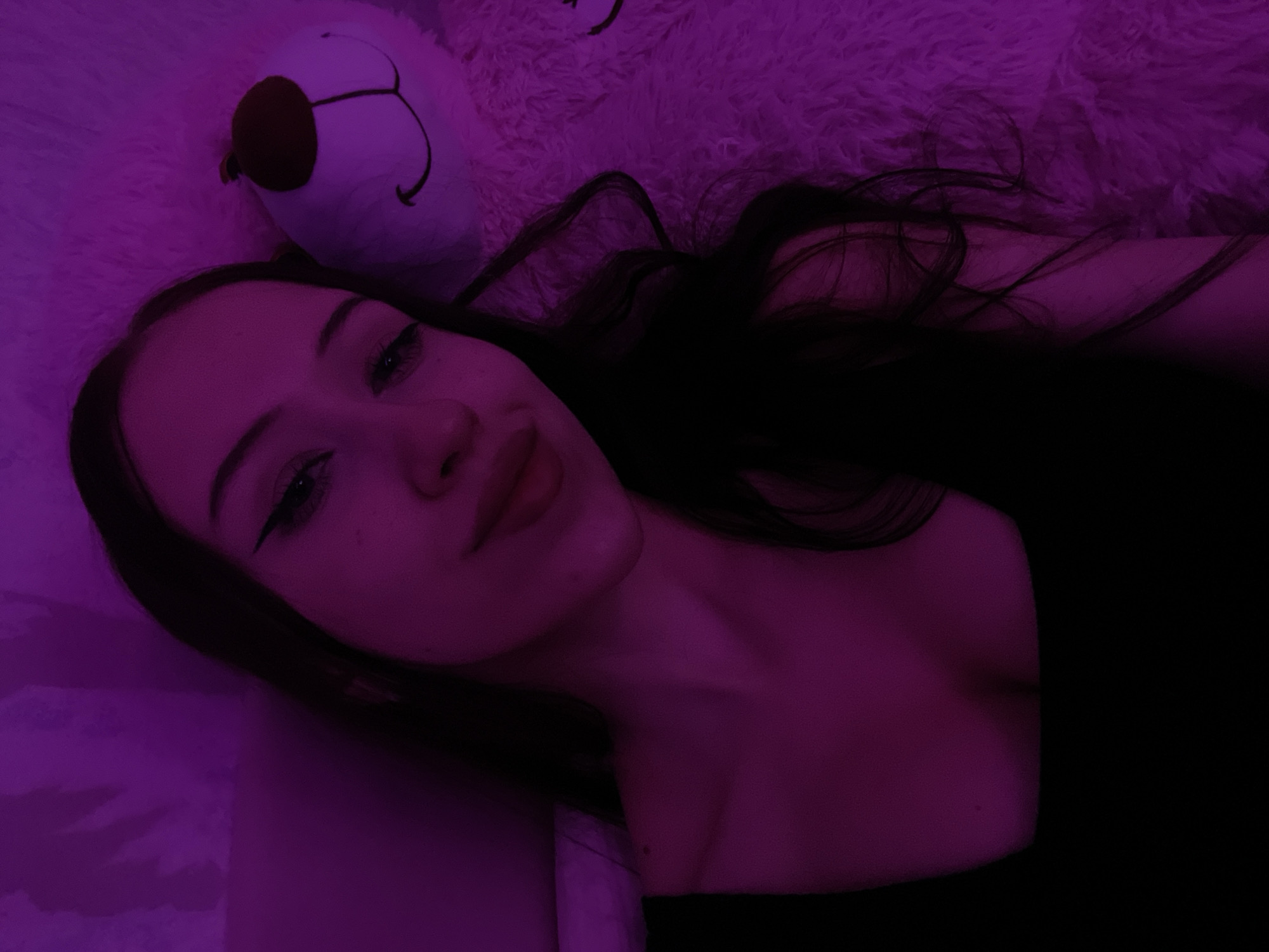 The image is a close-up selfie of a young woman taken in Votkinsk, Russia. She is lying down, possibly in bed, with a large, fluffy pink teddy bear beside her. The lighting is dim and casts a purple hue over the scene. The woman is looking at the camera with a slight smile. She is wearing a black top. The background is a textured surface, likely bedding.