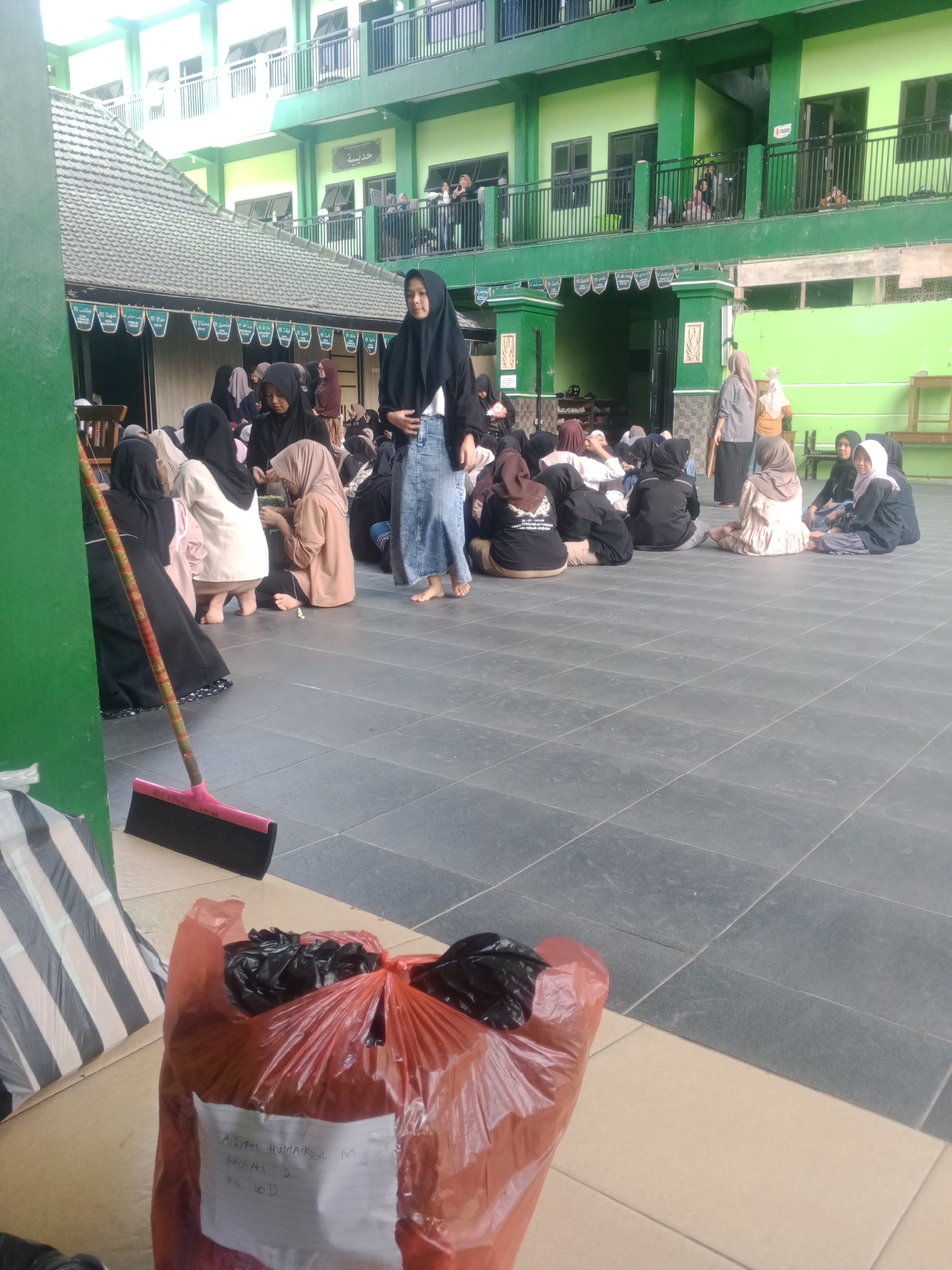 A group of young women in hijabs are seated on a tiled courtyard in front of a green and white building. The building appears to be a school or religious institution, with multiple levels and balconies. Some of the women are interacting, while others are looking around or resting. A broom is leaning against a green wall in the foreground, and a red plastic bag filled with trash is on the ground. The scene is illuminated by natural light, suggesting it is daytime. The architecture and the attire of the women indicate a location in Indonesia, potentially a Madrasah in Banyudono.