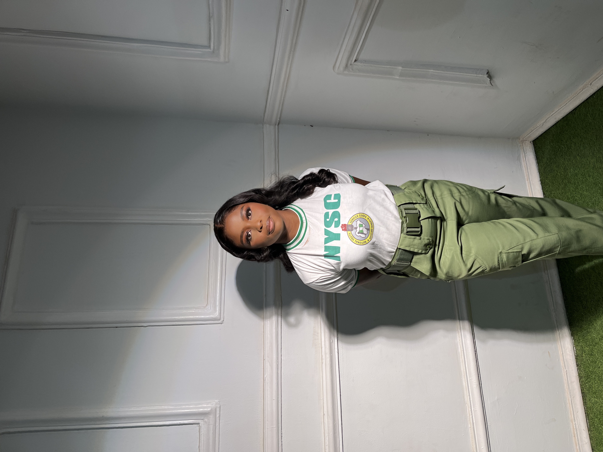 A young woman stands in the corner of a room. She wears a white t-shirt with green trim, and the acronym "NYSC" is prominently displayed on her shirt. Additionally, the t-shirt features a circular emblem indicating that she is part of the "National Youth Service Corps", in green, yellow, and white. Her trousers are green and she has a matching belt. The walls are a pale gray and have raised, framed paneling. The floor is covered in green, synthetic turf. The location in the city of Jos, Nigeria cannot be determined from the visual.