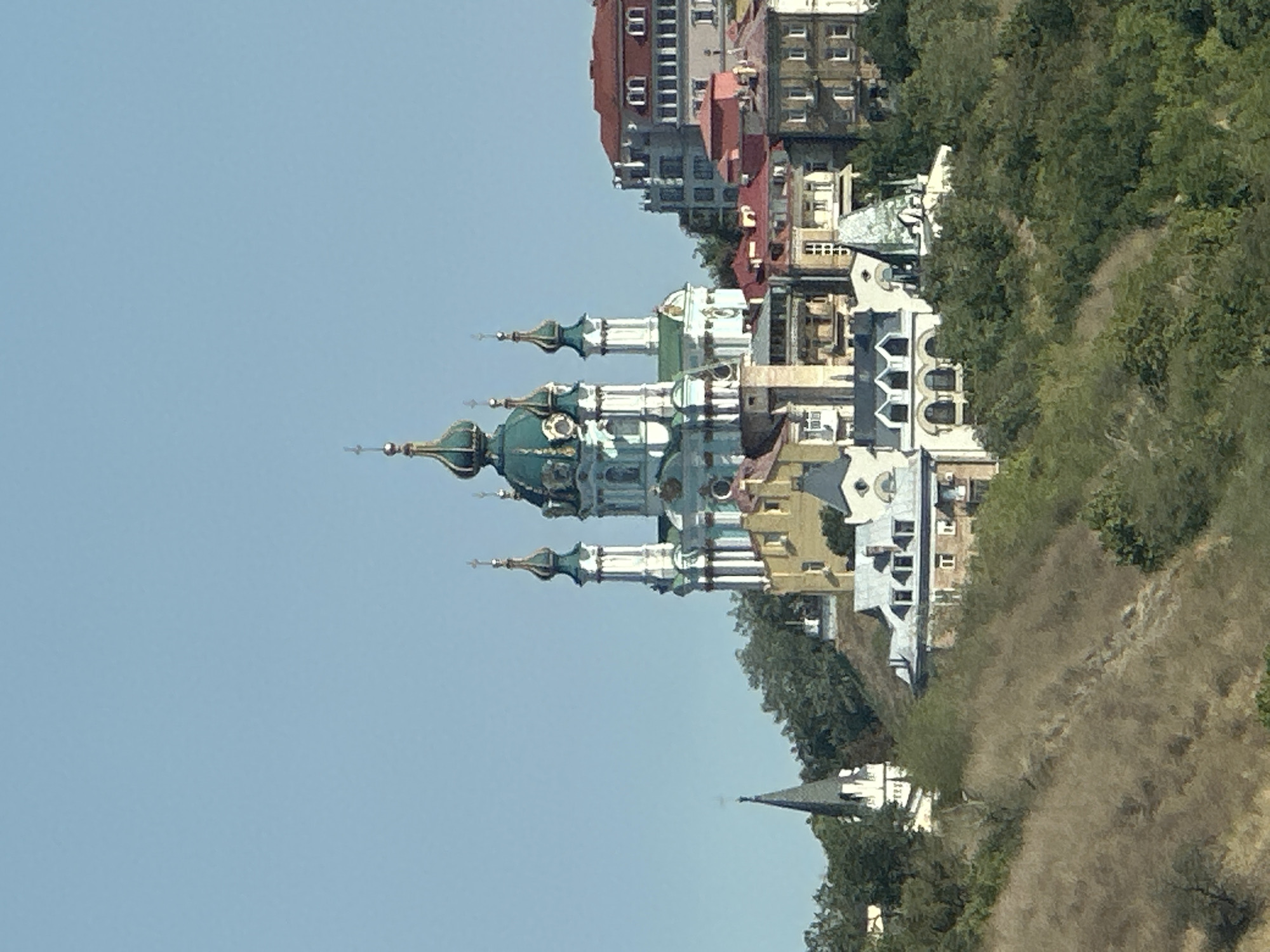 The image displays a panoramic view of St. Andrew's Church in Kyiv, Ukraine, situated on a hill. The iconic blue and white Baroque church with its green domes is prominent in the center of the frame. Surrounding the church are various other buildings, including residential structures and a tall, slender spire on the left. The foreground consists of a steep, dry, grassy slope, indicative of a hillside. The sky is a clear, light blue, suggesting a sunny day. The scene is captured from a distance, providing a wide view of the architectural ensemble and its natural surroundings.