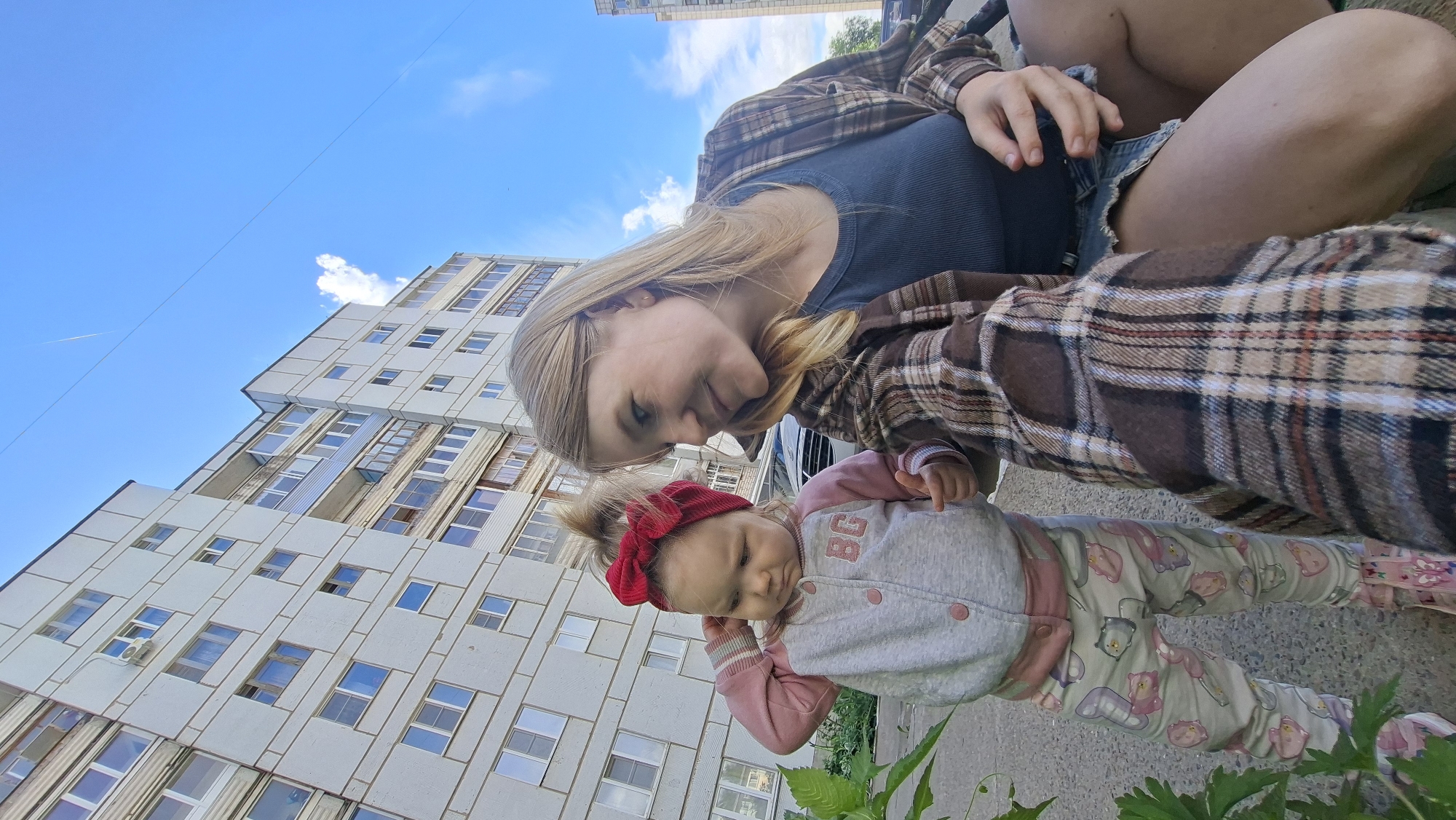 In Chaikovsky, Russia, on a sunny day, a young woman and a toddler are positioned in front of a light-colored, multi-story apartment building. The woman, wearing a plaid shirt over a dark tank top and a denim skirt, is crouching and looking down at the toddler. The toddler, with a red bow in her hair, is wearing a light gray and pink jacket with buttons and patterned leggings. The background shows a clear blue sky with a few wispy clouds and a contrail from an airplane. There is a green plant in the foreground to the left of the frame.