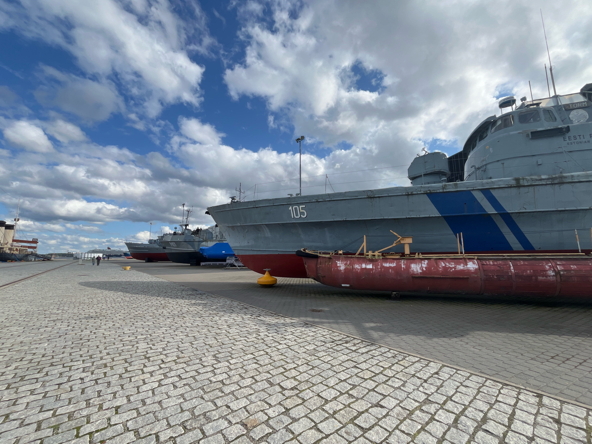 In Tallinn, Estonia, a line of decommissioned naval ships rests on dry land within a harbor area. The foreground is dominated by a cobblestone path, leading the eye towards a row of gray vessels. The largest ship in the foreground displays the number "105" on its hull and features a distinctive blue stripe. A smaller red hull of another vessel is visible beneath the main ship. Further down the line, additional ships are visible, one covered with a blue tarp. In the background, a larger ship with a white superstructure is docked. Two individuals are walking on the paved area between the cobblestones and the ships. The sky above is a mix of blue and scattered white clouds.