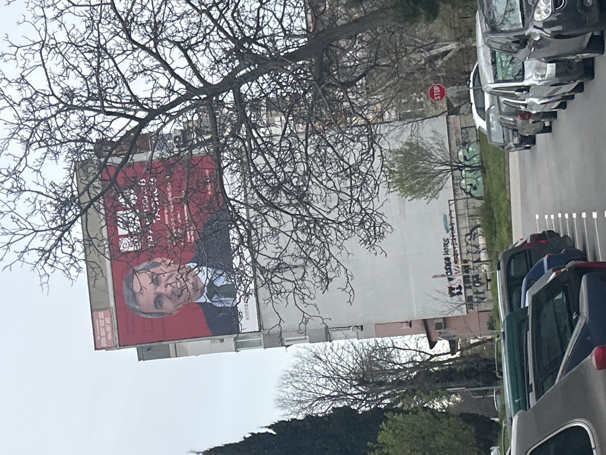 The image shows a large billboard on the side of a building in Varna, Bulgaria. The billboard features a portrait of a man in a suit against a red background. Part of the billboard and building is obscured by a bare tree in the foreground. To the right of the building, several cars are parked along a curb. Graffiti is visible on a wall adjacent to the parked cars. A "STOP" sign is also visible. The sky is overcast. The scene suggests a somewhat urban environment with elements of commercial advertising and public infrastructure.