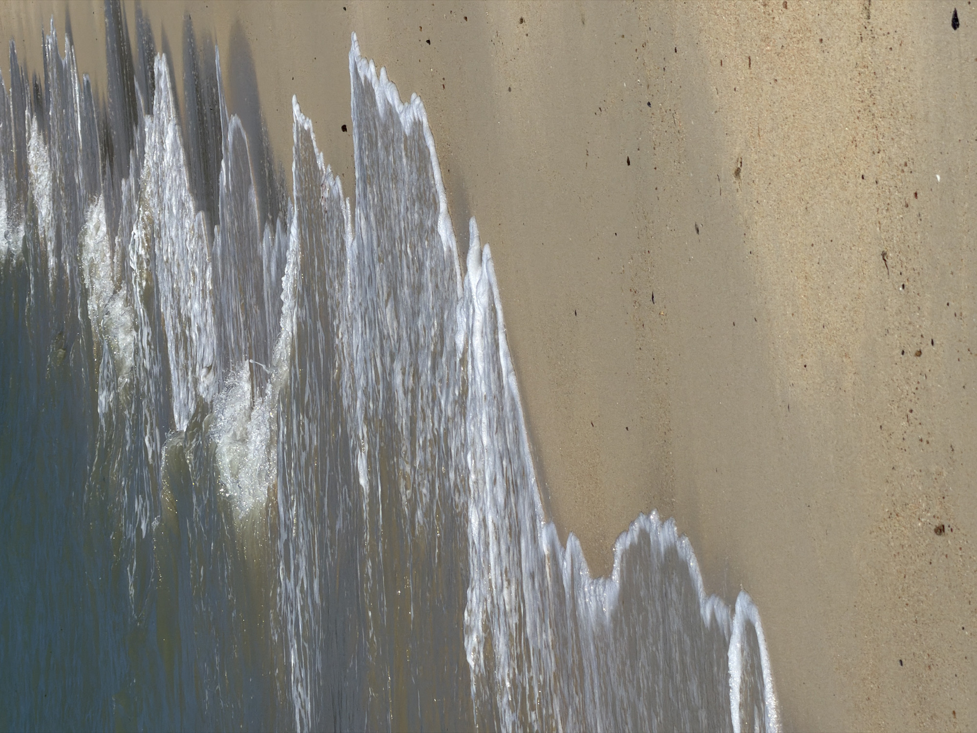 The image shows a close-up view of waves gently lapping onto a sandy beach. The water is a murky green-brown, and small white foam crests are visible on the waves as they break. The sand is light tan and appears to be composed of fine grains with occasional small, dark specks that could be shells or debris. The perspective is from a high angle looking down at the shoreline. No people or man-made structures are visible in the frame, suggesting a natural coastal environment. The image is likely taken in Ko Samui District, Thailand, which is known for its beaches and coastal scenery.