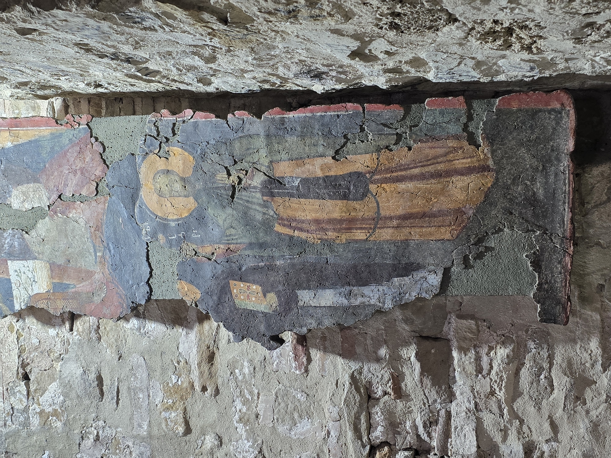 The image depicts a fragment of a fresco, likely of religious iconography, adhered to a rough-hewn stone wall. The fresco is heavily damaged with cracks, missing pieces, and surface degradation, revealing the underlying material in several areas. Coloration is subdued, featuring blues, yellows, browns, and reds. The style suggests Byzantine or a related Eastern Christian artistic tradition. Given the nature of the scene, it is possibly located in an archaeological site, a historical building, or religious institution. The location is noted as Unknown, Unknown.