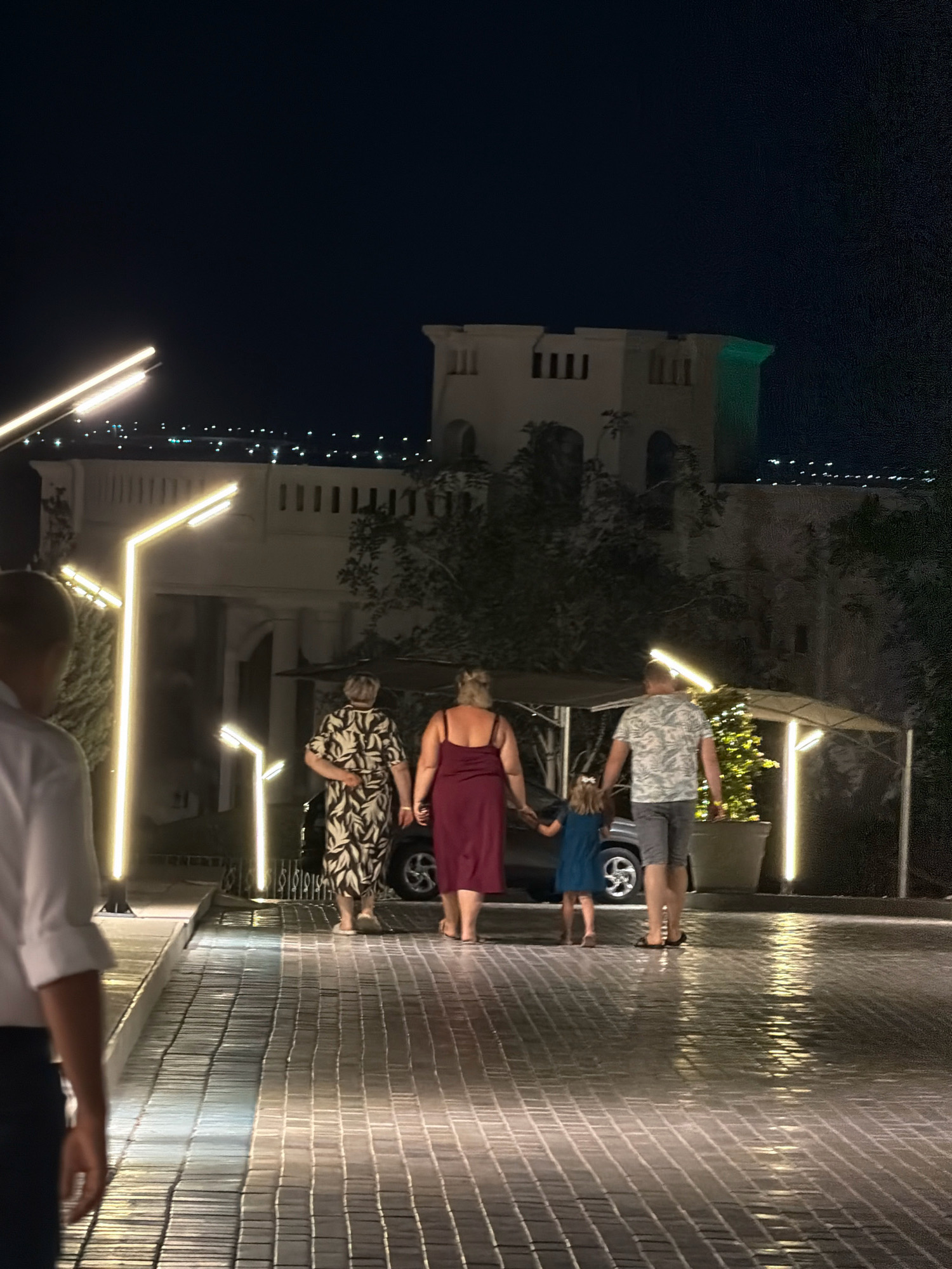At night in Sharm El Sheikh, Egypt, a family walks down a tiled walkway towards a building with illuminated architectural features. The walkway is lined with modern, vertical light fixtures that cast a warm glow. In the background, a large building with a pale facade and darker archways is visible, illuminated by lights and a green light on its upper section. The distant cityscape of Sharm El Sheikh, with its numerous small lights, is seen against the dark sky. The family consists of two adults and a young girl, all walking away from the viewer. One adult woman is wearing a patterned dress, another is in a maroon dress, and the man is in a patterned shirt and shorts. The young girl is holding the hand of the man and the woman in the maroon dress. A car is parked partially under a canopy behind the family. The walkway's tiles are wet and reflect the surrounding lights, suggesting recent rain or maintenance. Lush vegetation is present to the right and behind the building.