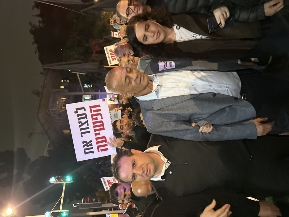 A group of people is gathered at night. Several are holding signs, one of which reads in Hebrew to "Stop the Crime." There are also signs stating "Arab Lives Matter" and "Stop". The crowd appears to be at a protest or rally. A man in a black sweater is looking directly at the camera, with a group of people surrounding him. A man in a grey suit and a woman in a brown jacket are standing beside him.