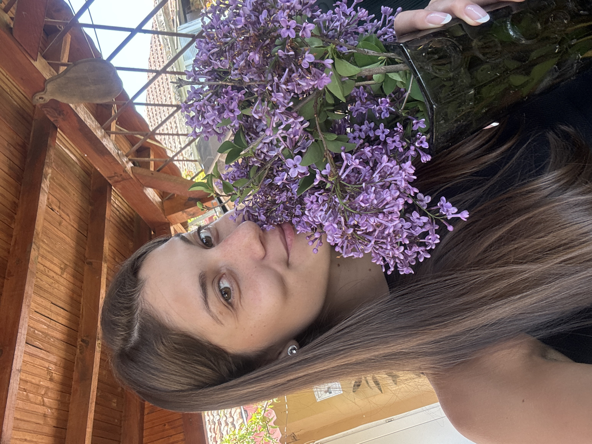 A woman with long brown hair is holding a bouquet of purple lilacs in front of her face. She is standing under a wooden structure. In the background, there is a building. The location is Rusokastro, Bulgaria.