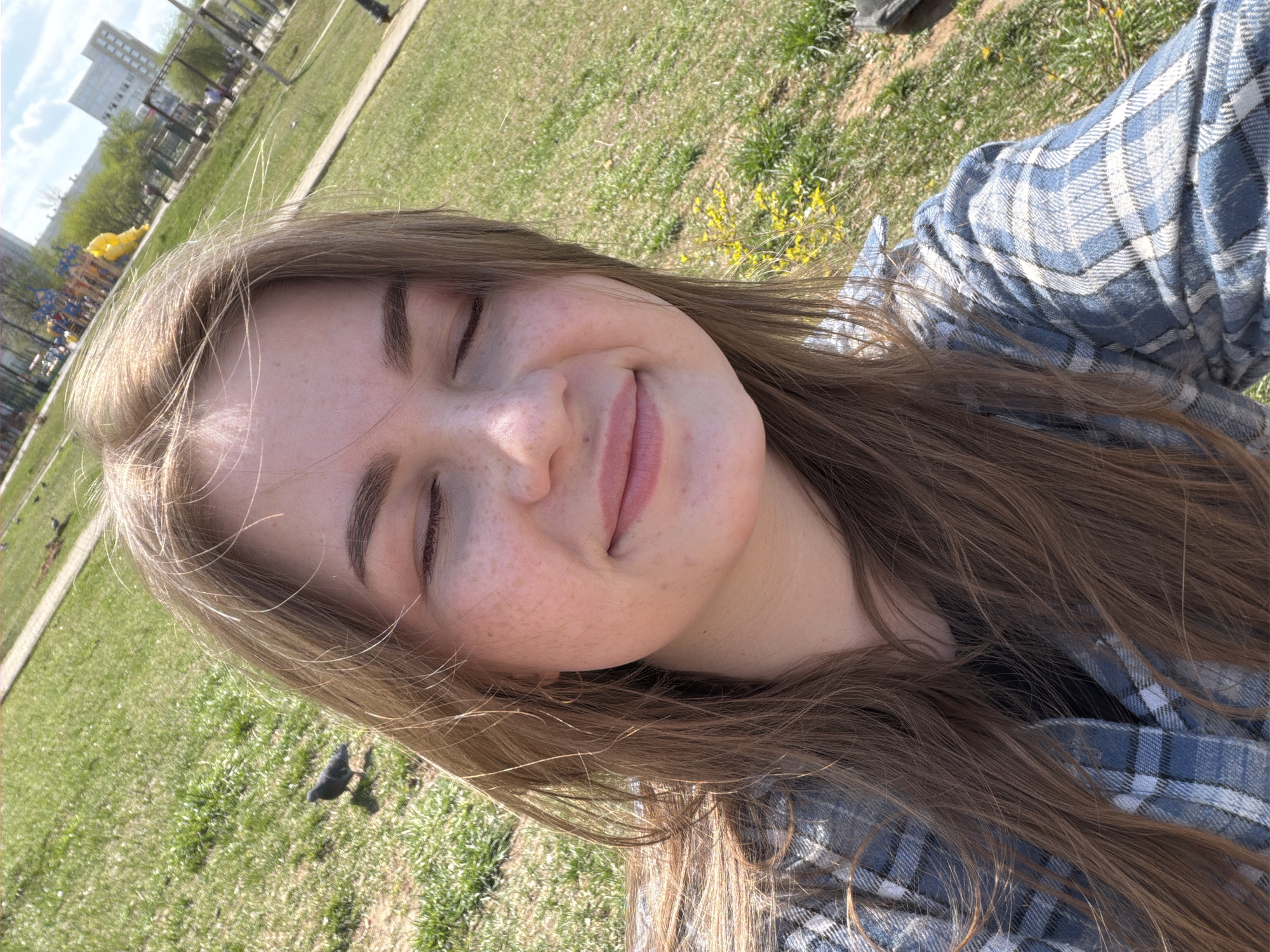 A young woman is photographed outdoors, likely in a park, on a sunny day. She has light brown hair, freckles, and is wearing a blue and white plaid shirt. Her eyes are closed, and she is smiling slightly. In the background, there is green grass, some trees, a playground with yellow structures, and a building with a red facade. The location is most likely Chaikovsky, Russia. A bird, possibly a pigeon, is on the grass at the lower left corner of the image.