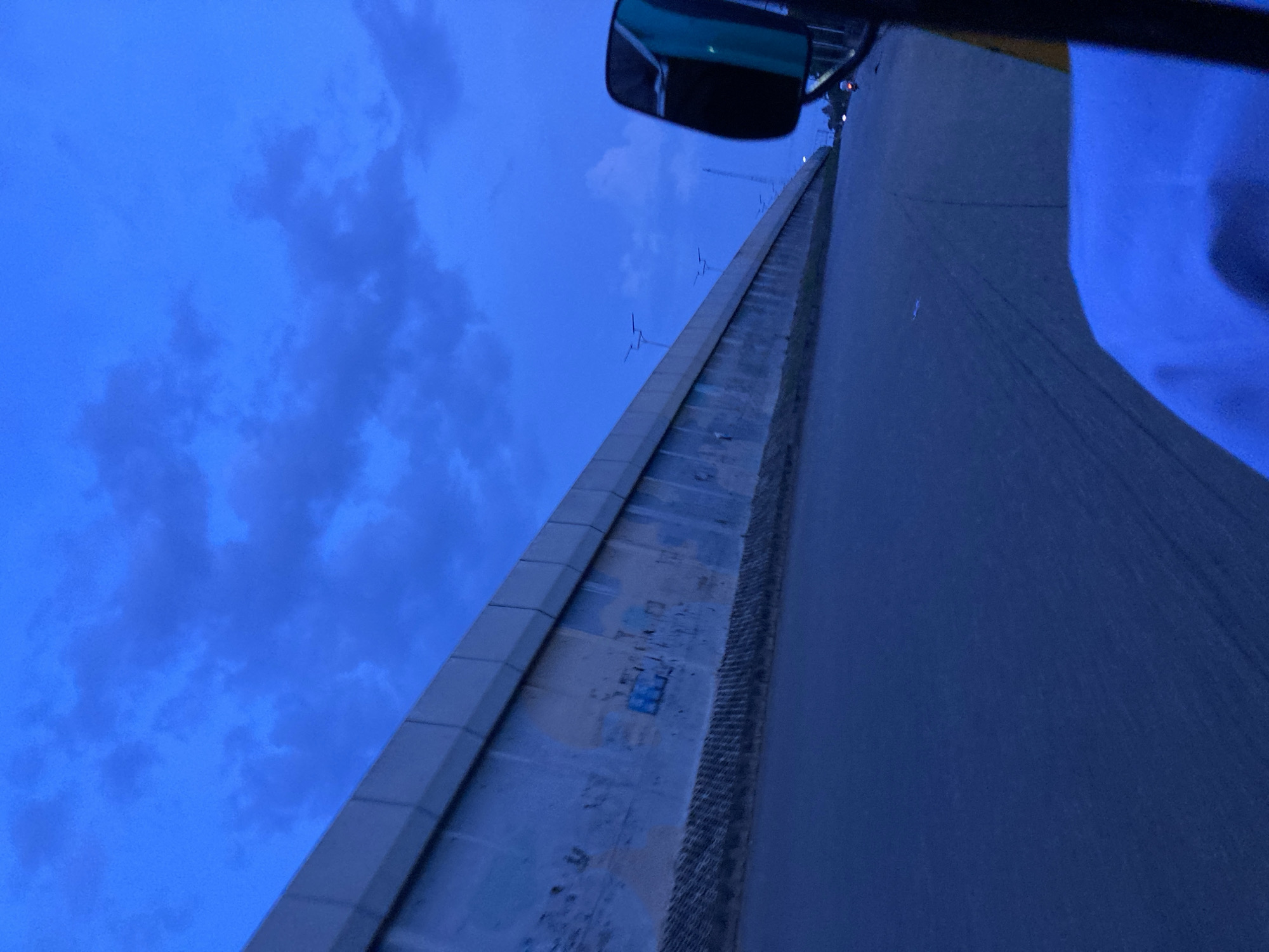 The image captures a view from a vehicle, likely a car or tuk-tuk, traveling on a paved road at dusk in Jos, Nigeria. A tall, concrete wall, possibly a barrier or part of an overpass, runs along the left side of the road. The wall is weathered and shows signs of graffiti. The sky above is a deep blue, with scattered clouds and streetlights visible in the distance. A side mirror of the vehicle is visible on the right, reflecting a portion of the sky and what appears to be a motorcycle with its lights on, moving away. The road surface is dark asphalt, with faint lane markings. The overall lighting suggests it is either early morning or late evening. The scene is devoid of any specific landmarks that would pinpoint an exact location within Jos, but it represents a typical urban or suburban road environment during twilight hours.